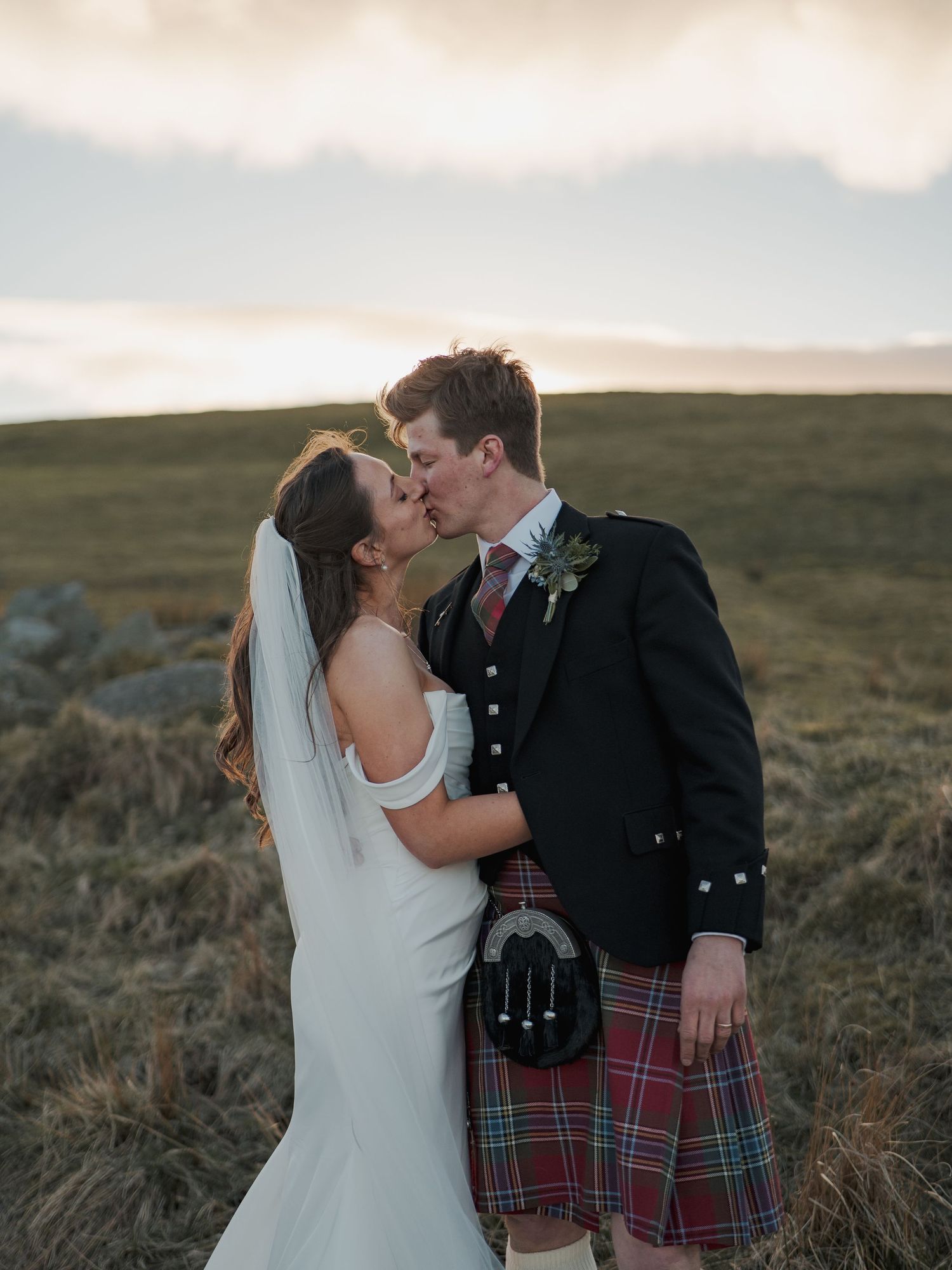 A couple shares a romantic kiss in a Scottish Highland setting, the groom wearing a traditional tartan kilt and the bride in a white gown.