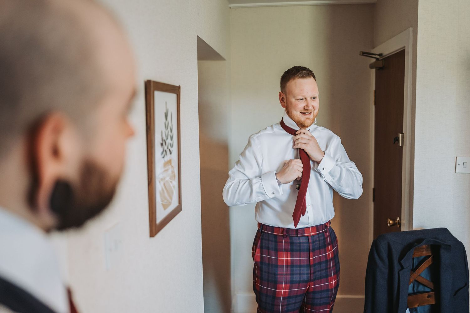 Groom adjusting red tie while wearing plaid kilt during wedding preparations at Oswald House.