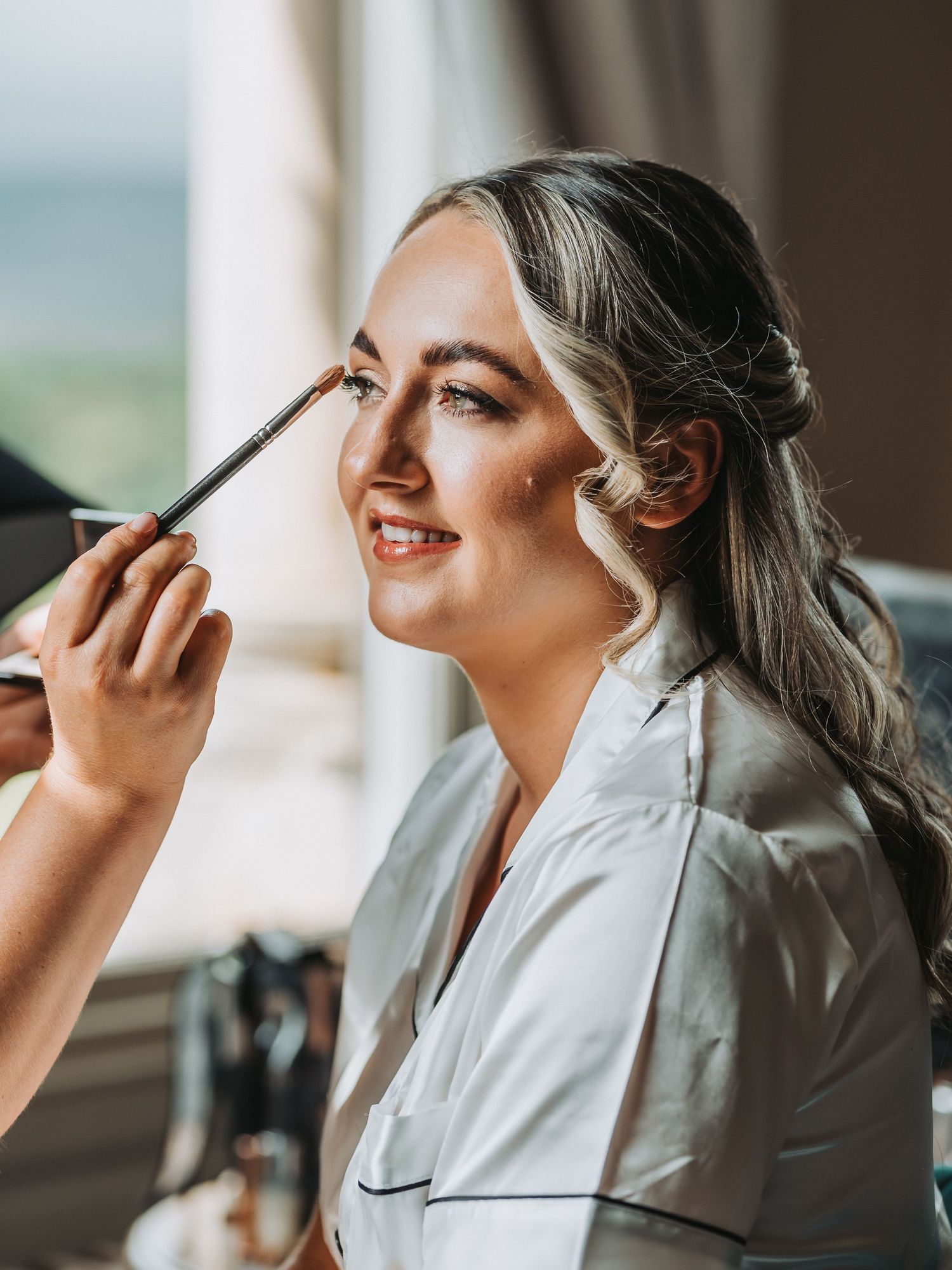 Bride getting makeup applied while seated by window during wedding preparation at Cornhill Castle.