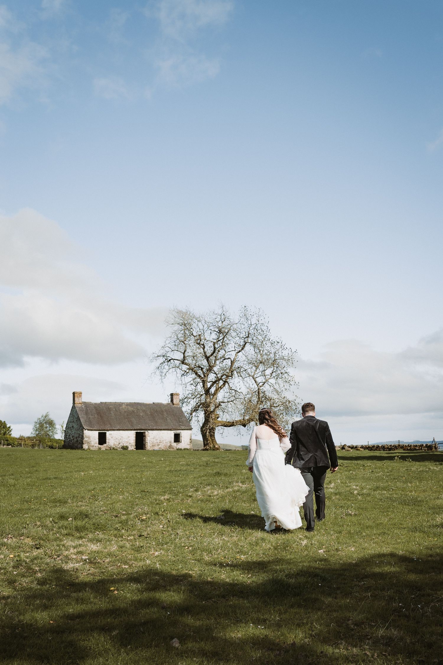 A couple walks across a green field towards a white bothy with a bare tree on a cloudy day in the countryside of Kinclune House.