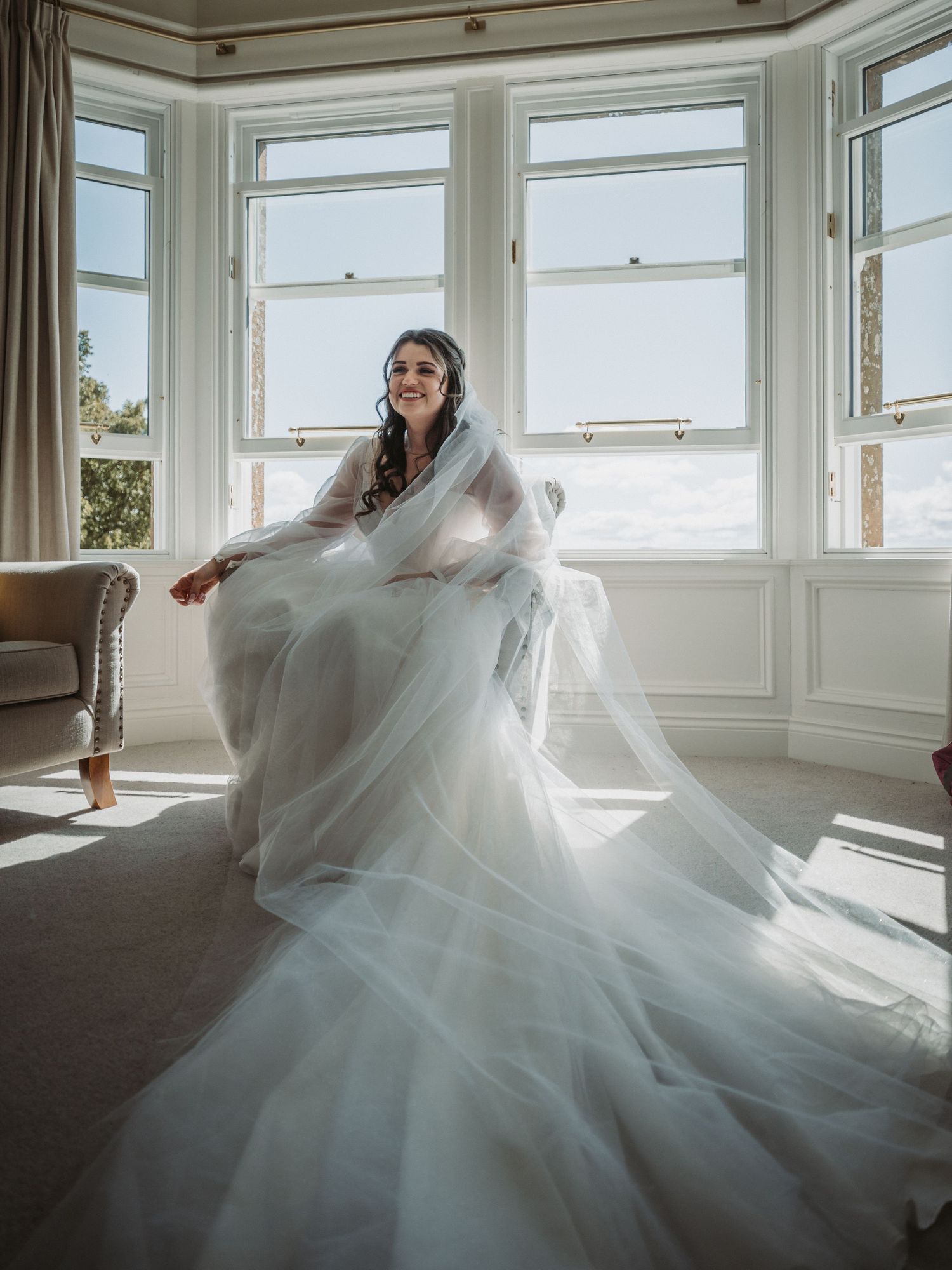 A person in an ethereal white wedding gown poses gracefully by a bright bay window in a sunlit room at Kinclune House.