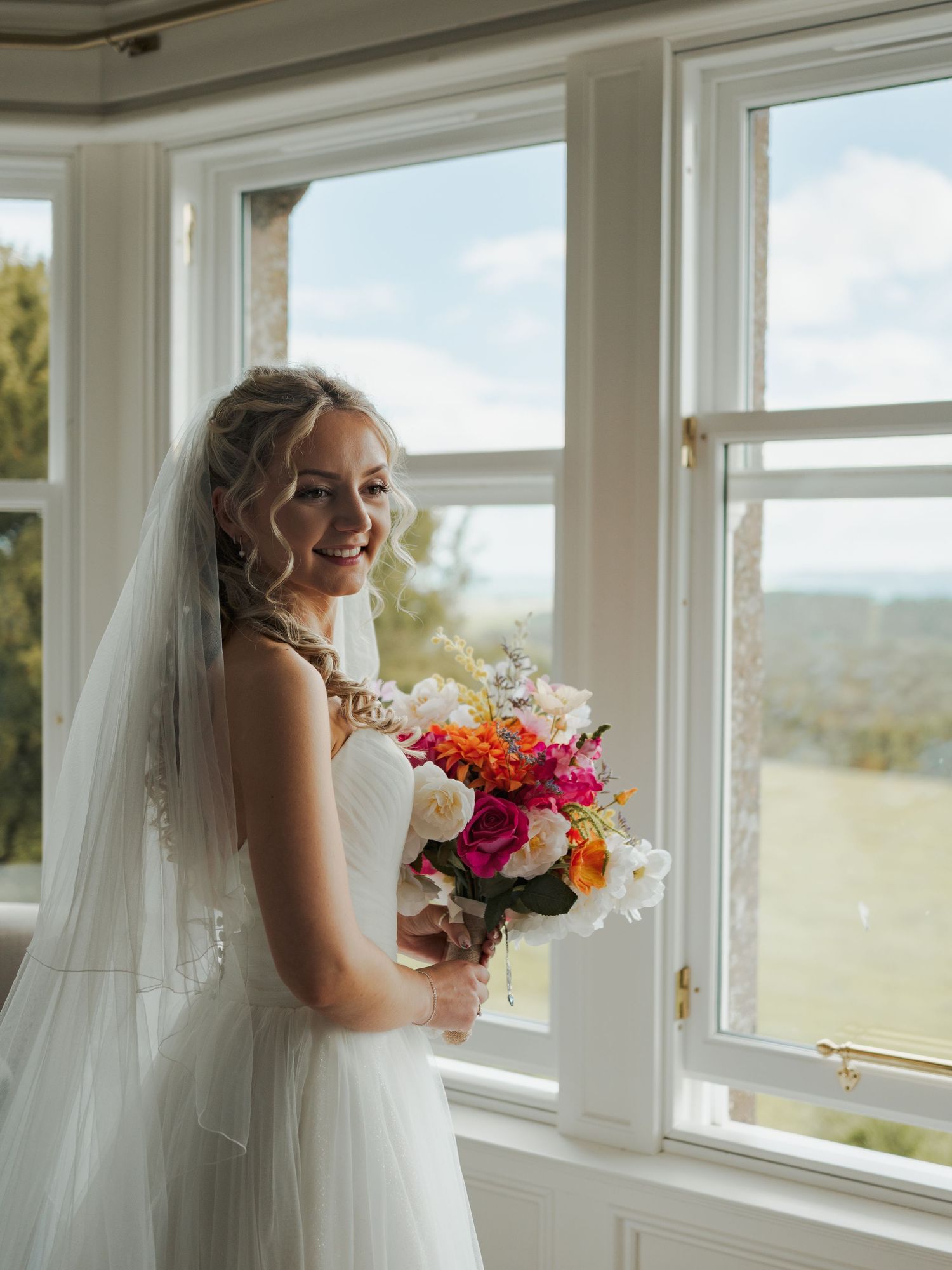 Smiling bride in white gown and veil holds colorful bouquet by sunlit windows with scenic countryside views.