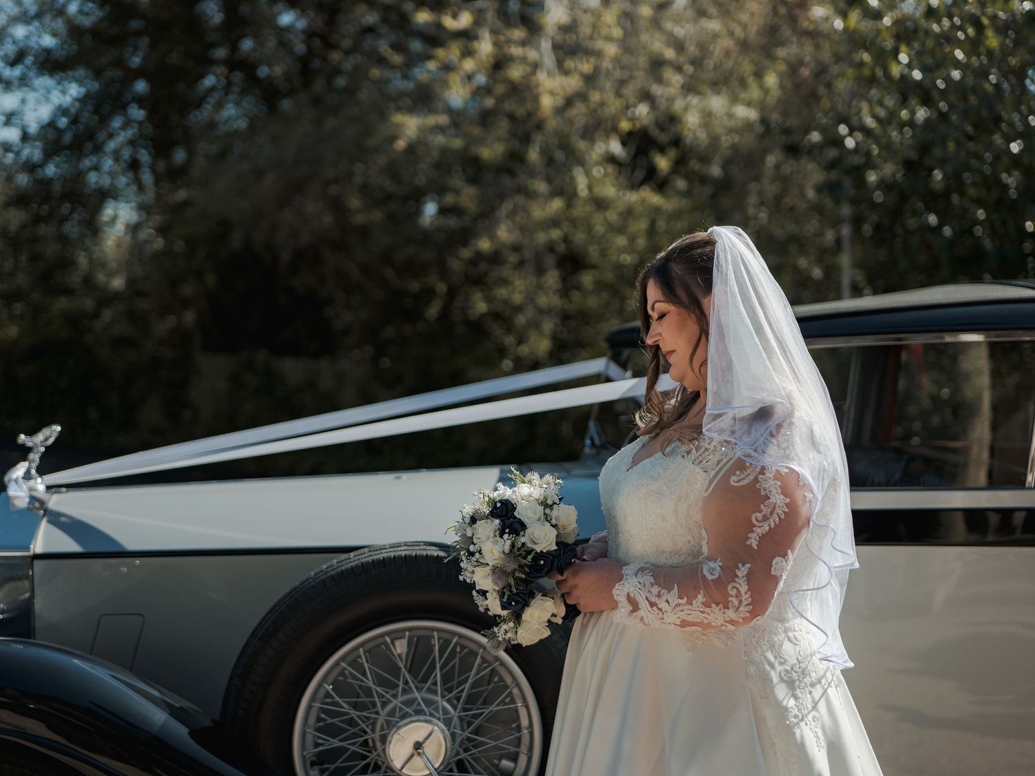 Bride in white gown and veil holds bouquet beside a classic vintage wedding car outdoors on a sunny day.