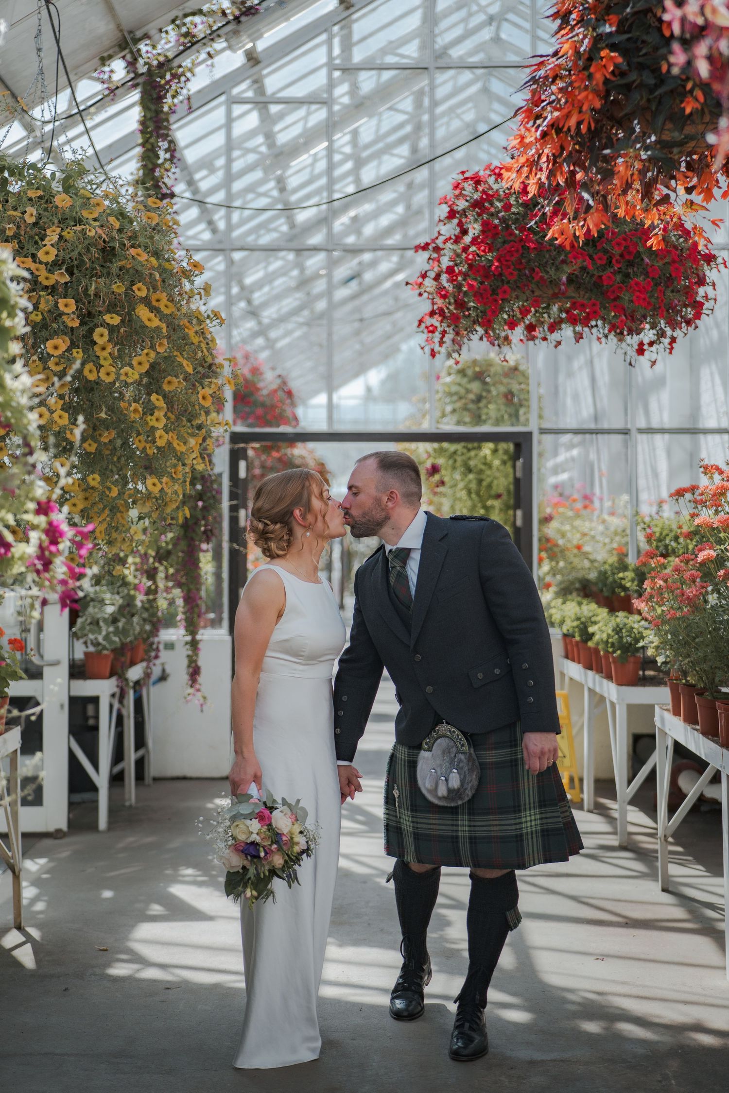 A couple in wedding attire sharing intimate moments in a greenhouse with hanging floral baskets and natural light.