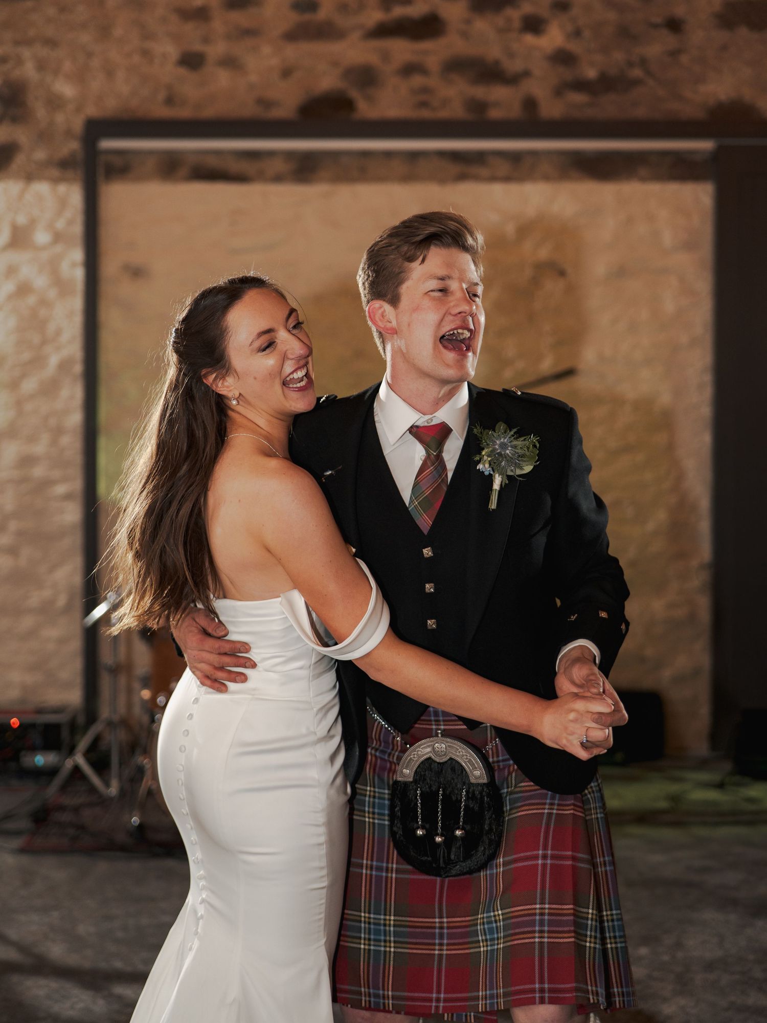 Bride in white gown and groom in Scottish tartan kilt laughing together during first dance at rustic wedding venue.