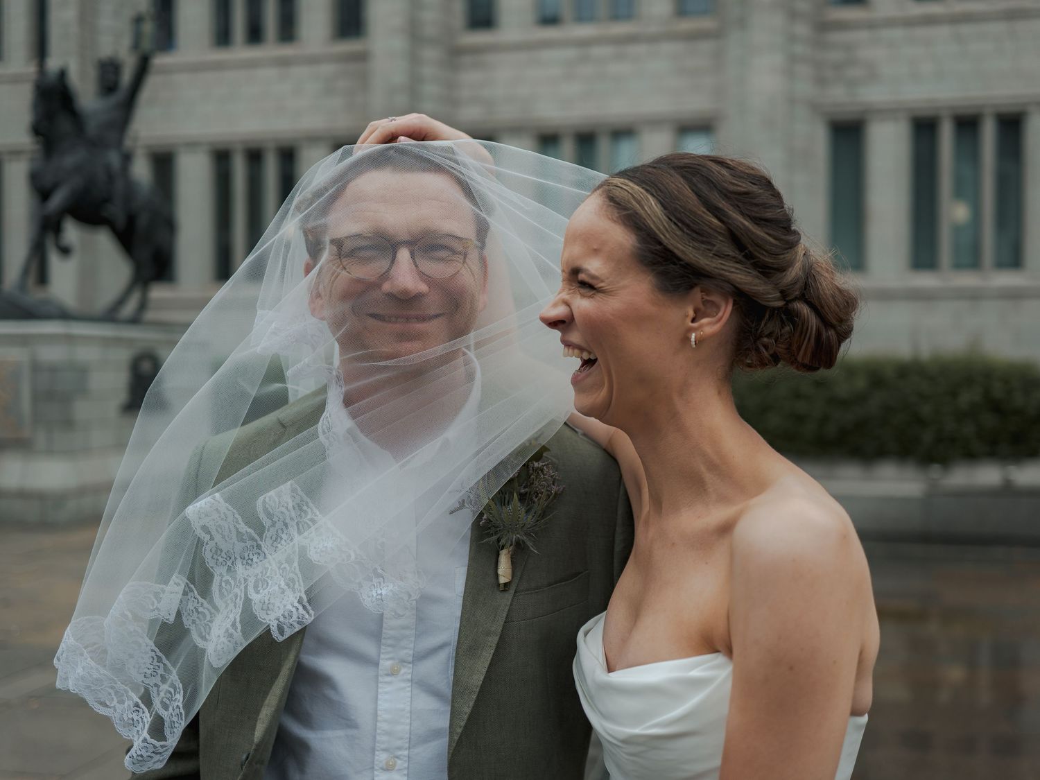 Bride and groom laughing together outdoors, veil blowing in wind, groom wearing tartan tie, urban monument background.
