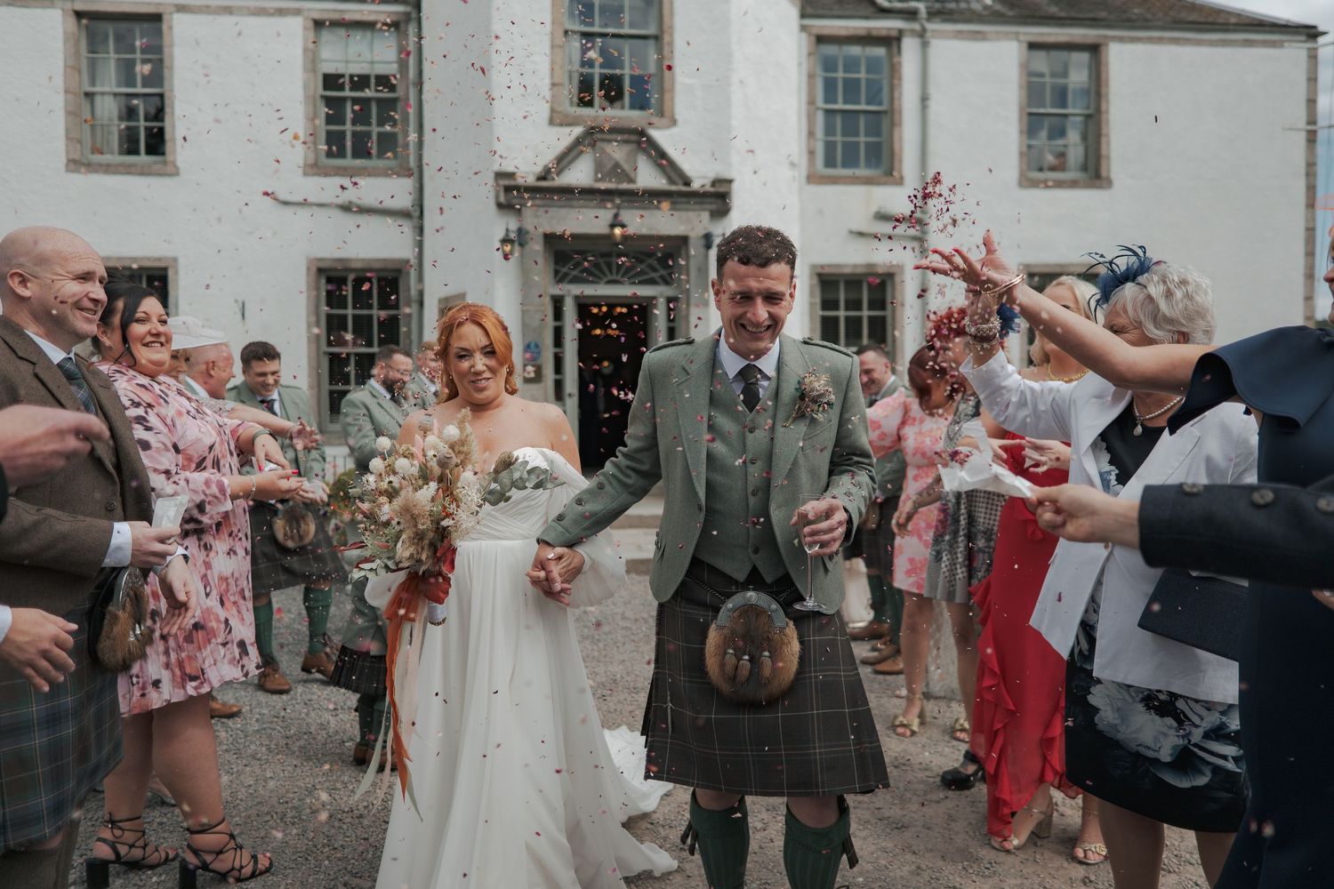 Wedding celebration outside a historic Scottish manor house as guests throw confetti on newlyweds in traditional attire at Banchory Lodge.