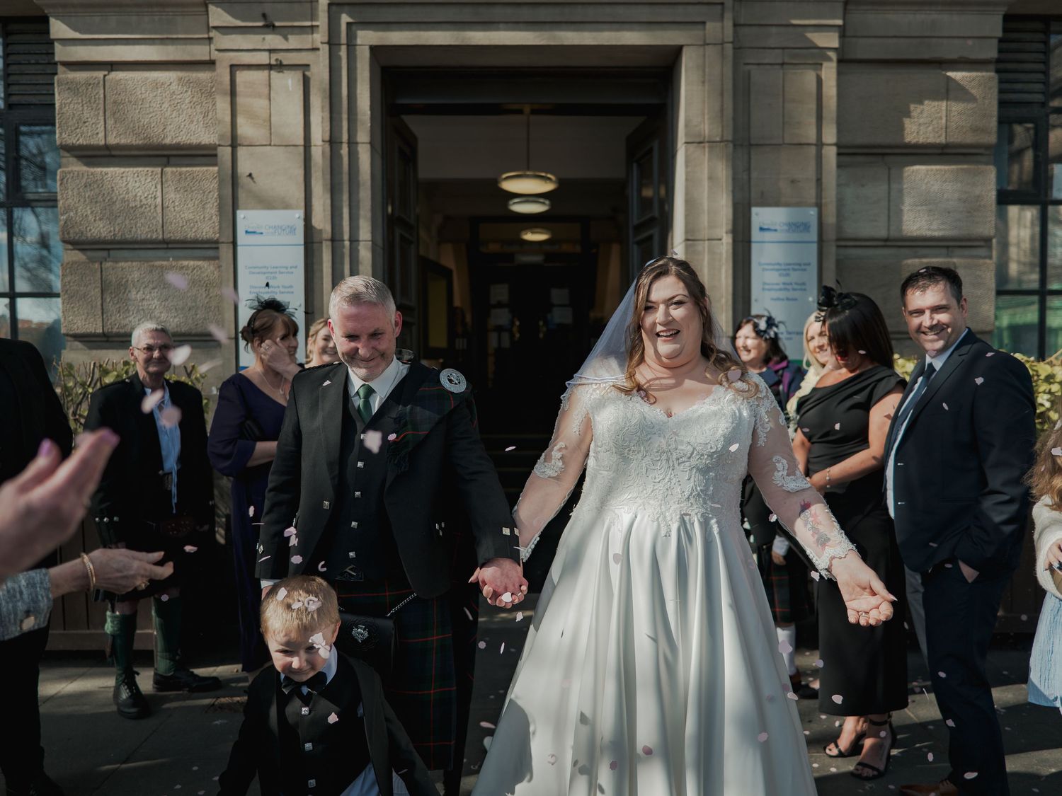 Bride and groom exit a building hand-in-hand with a child, guests throwing confetti in a joyful wedding celebration.