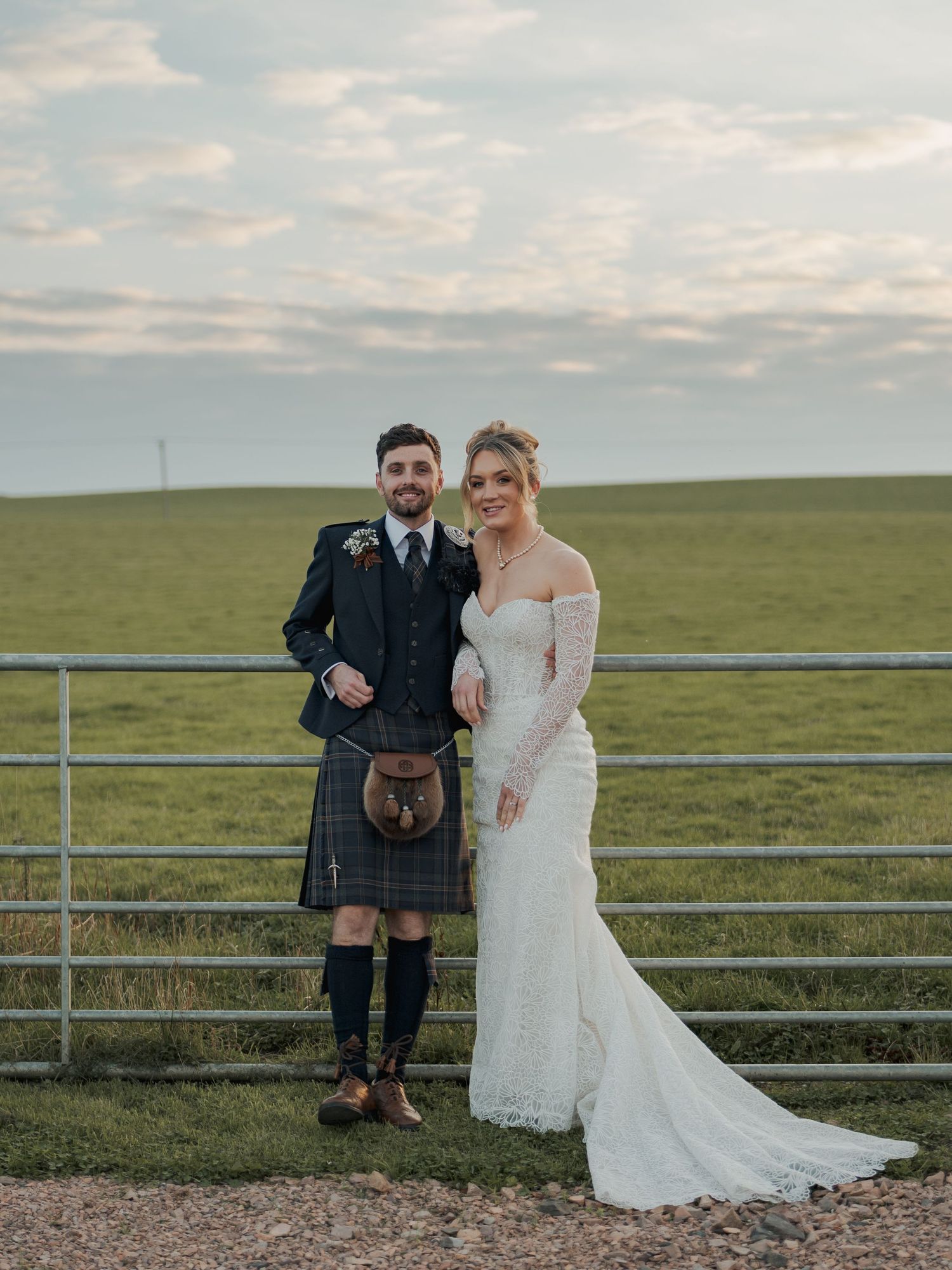 A couple poses together against metal fencing with rolling green hills and cloudy skies in the background at Falside Mill.