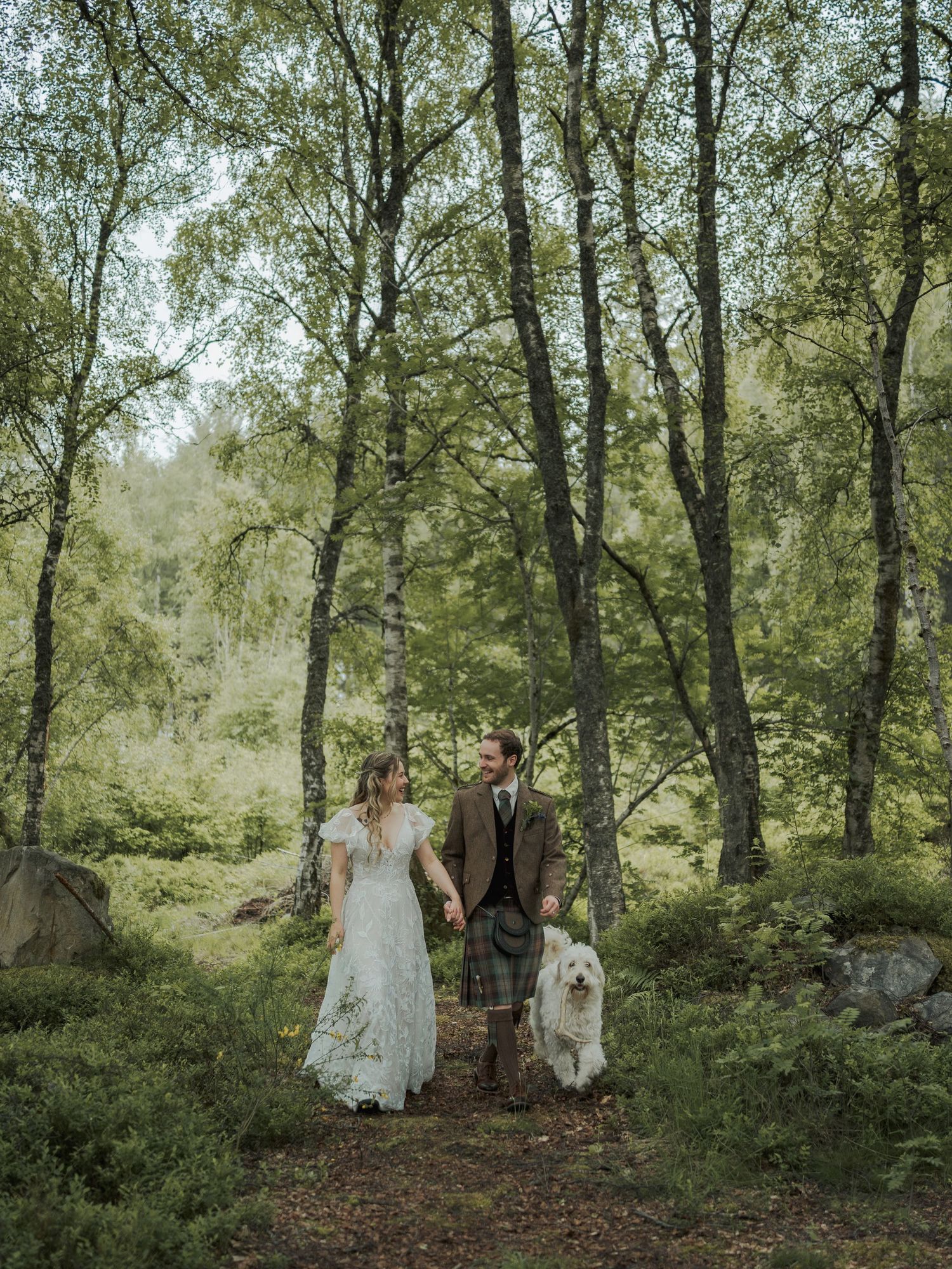 A bride and groom walk hand-in-hand with a white fluffy dog through a lush green woodland forest in Scotland.