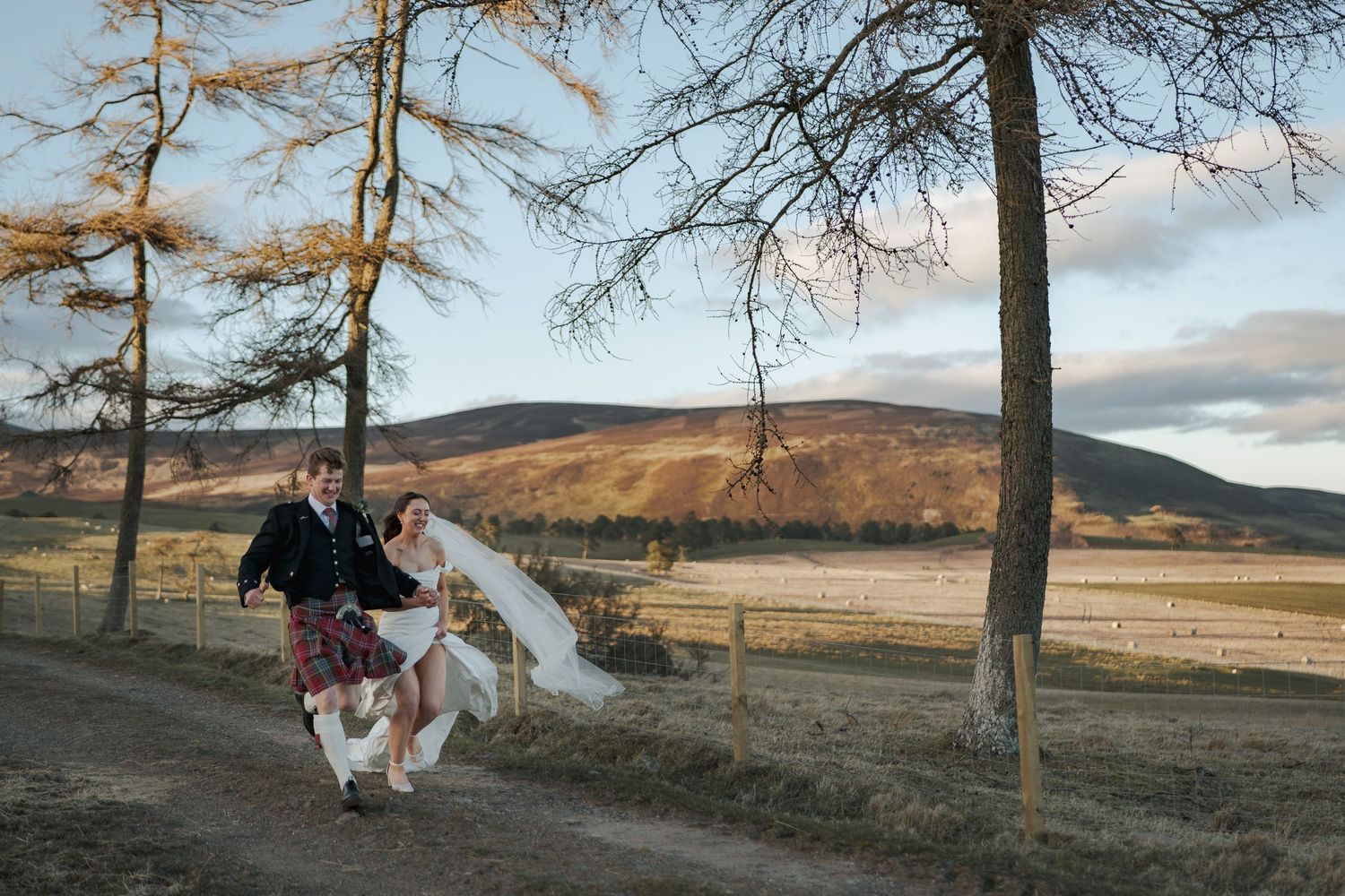 Bride and groom run along a farm track with stunning Kirriemuir countryside in the background at sunset