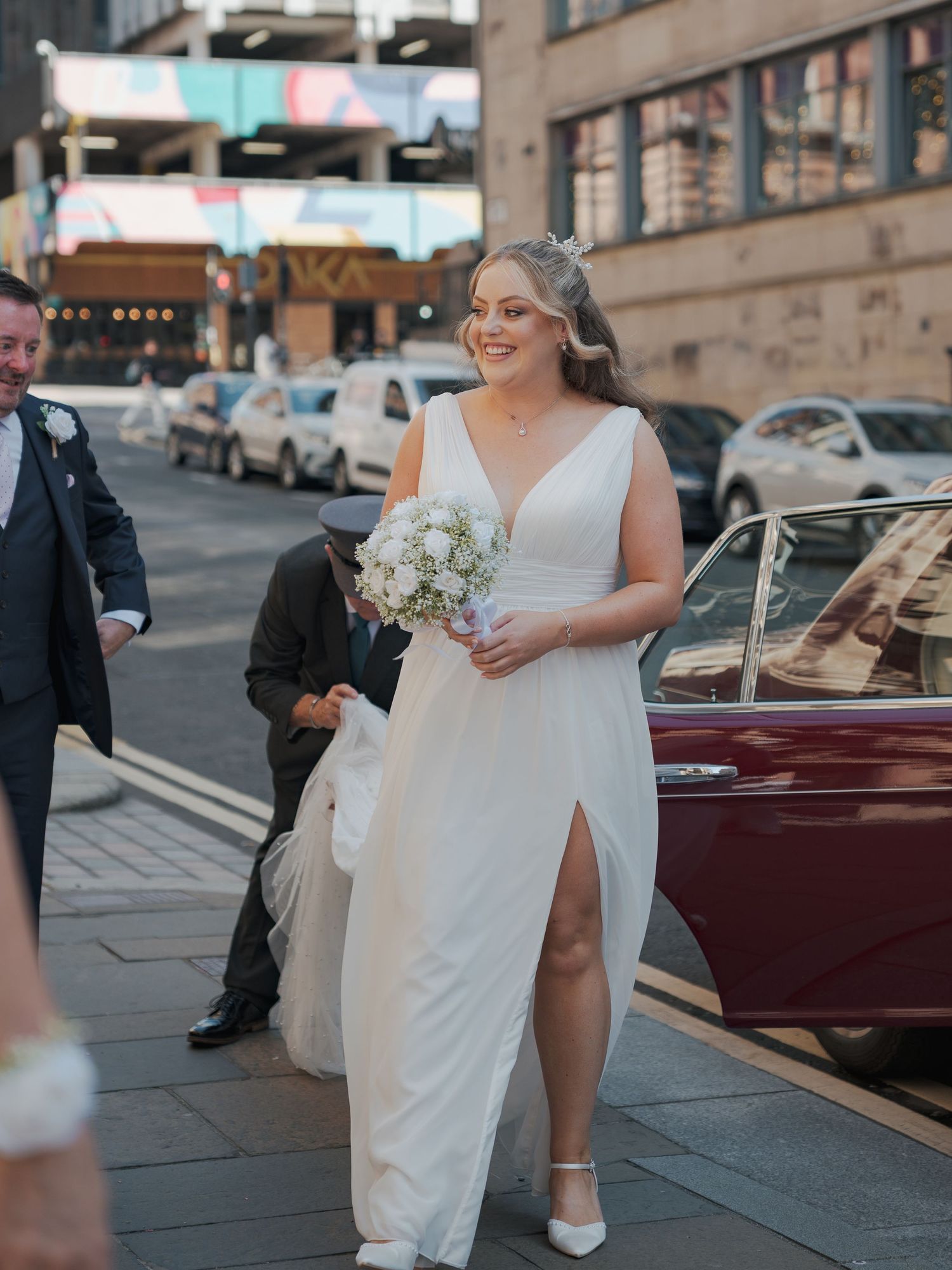 A bride in a white wedding dress stands beside a vintage red car outside 23 Montrose Street.