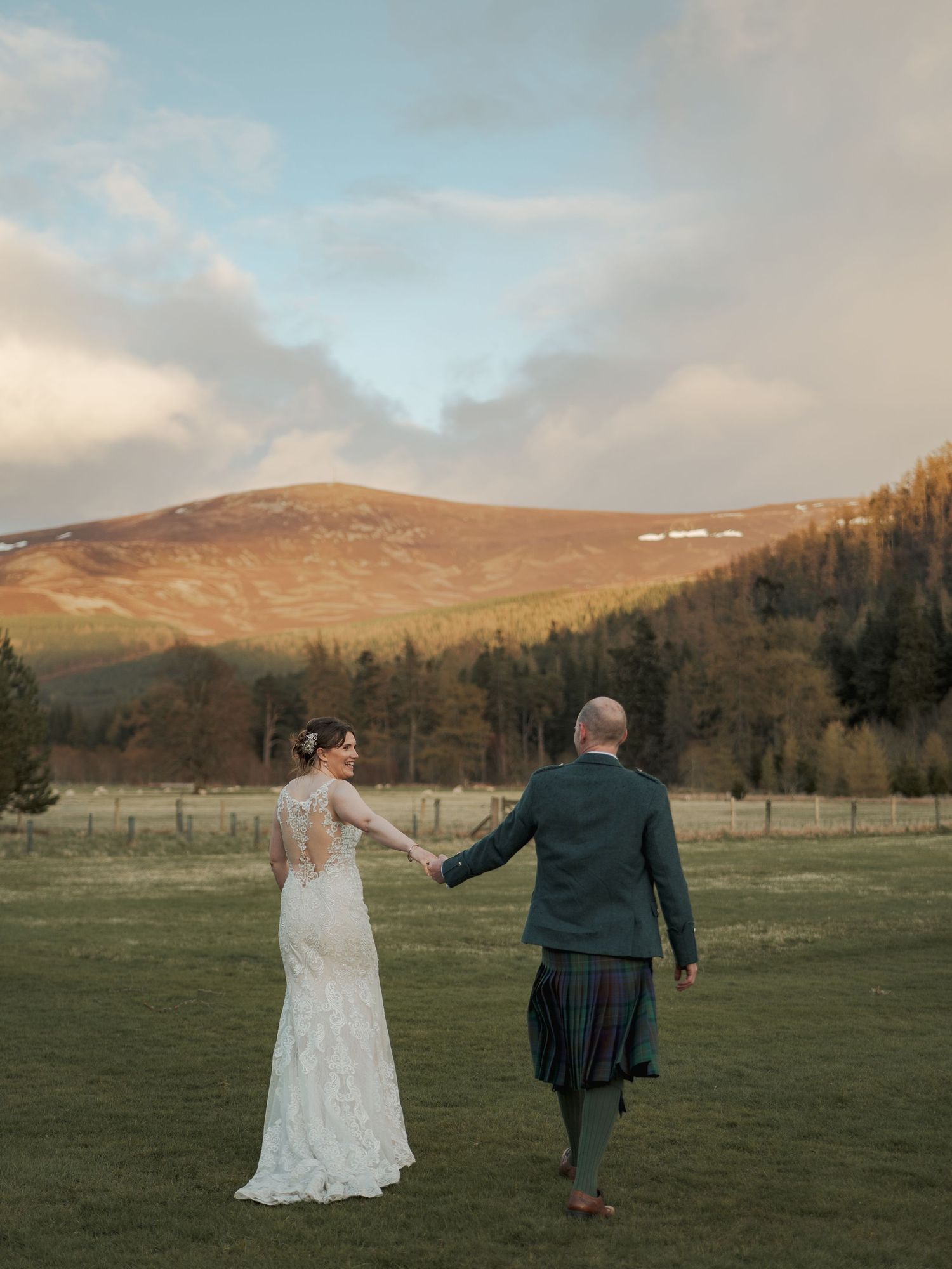 Bride and groom holding hands in Scottish Highland landscape at golden hour, with mountain backdrop and pine forest.