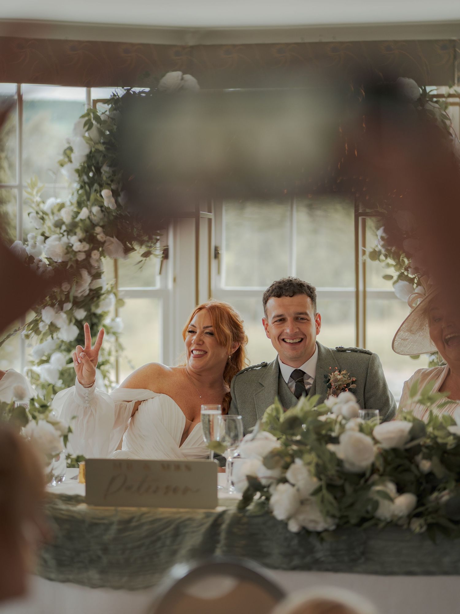 A joyful wedding celebration moment captured at the sweetheart table under an elegant floral arch with white roses.