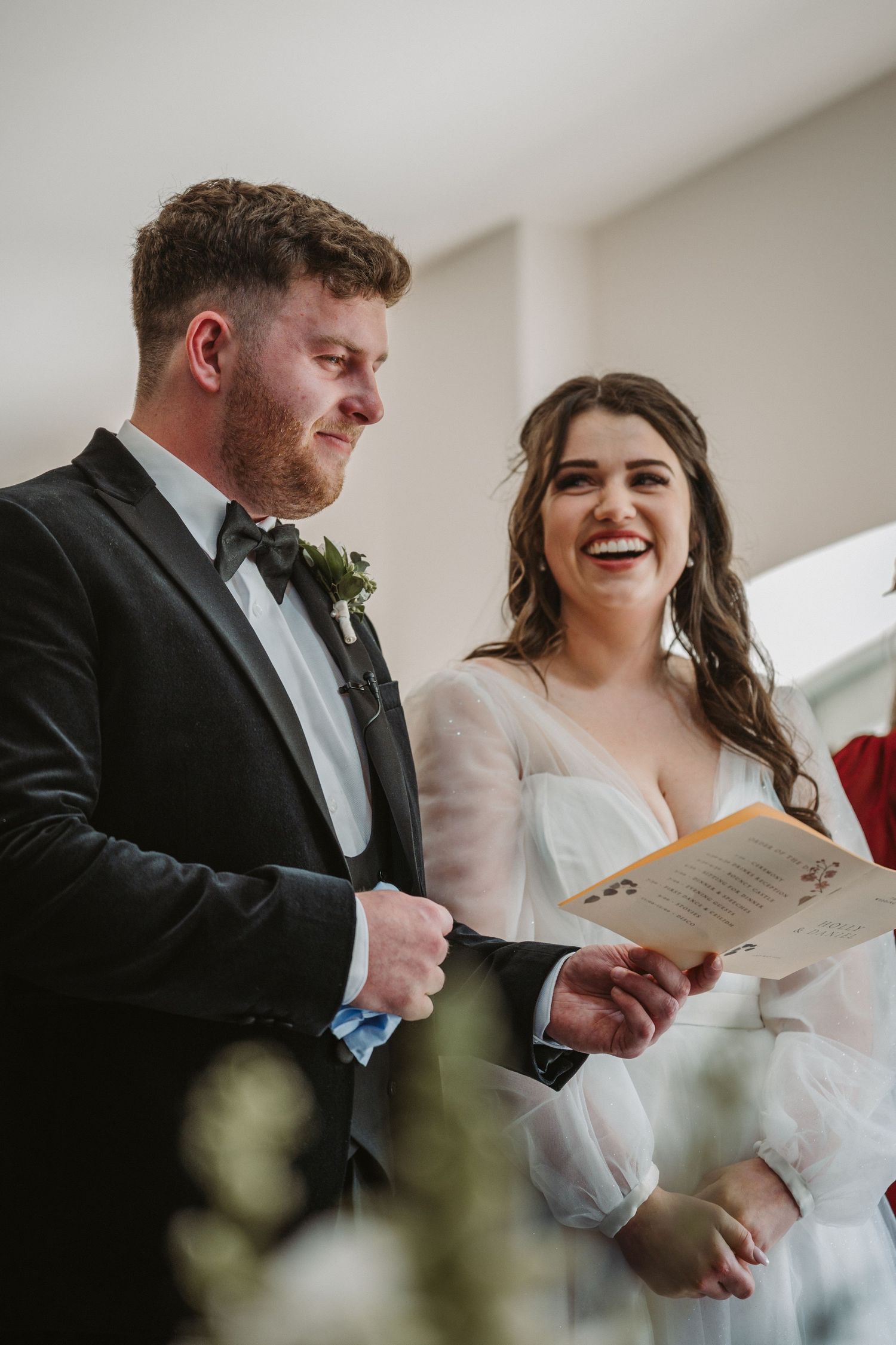 A couple laughs together during their wedding ceremony while holding ceremony programs in a bright indoor venue.