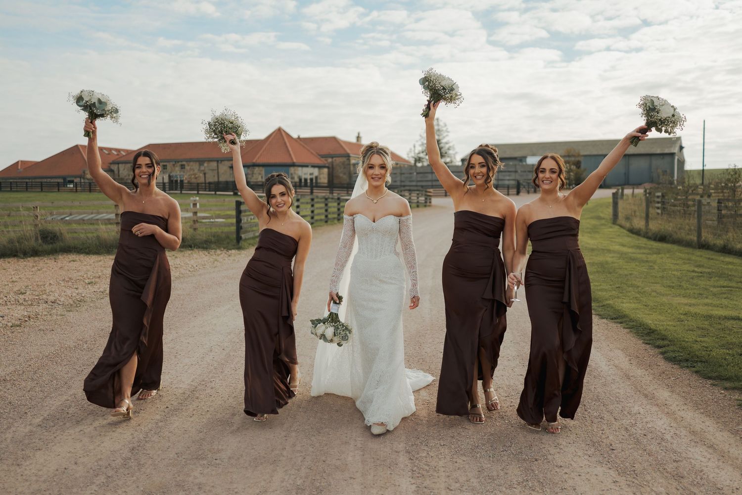 Bride in white gown walks with four bridesmaids in dark brown dresses raising bouquets on a rustic gravel path.