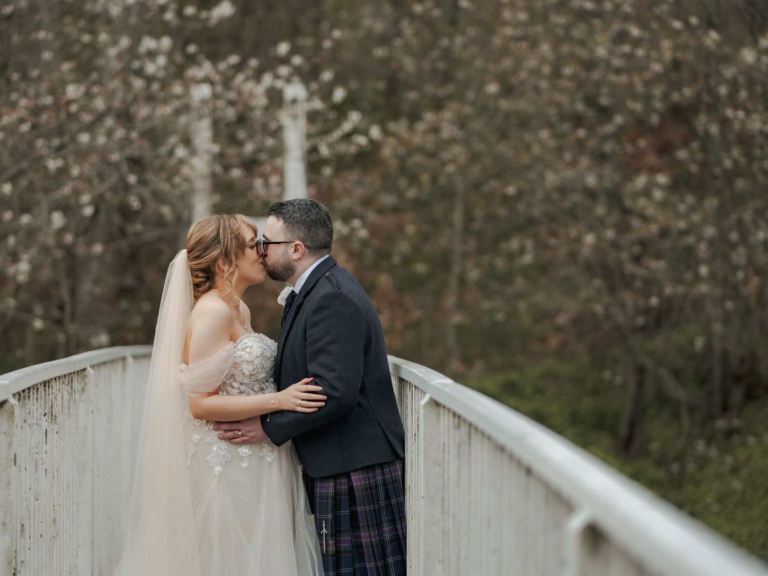 Bride and groom share a romantic kiss on a white bridge surrounded by blooming trees in a scenic outdoor wedding photo.