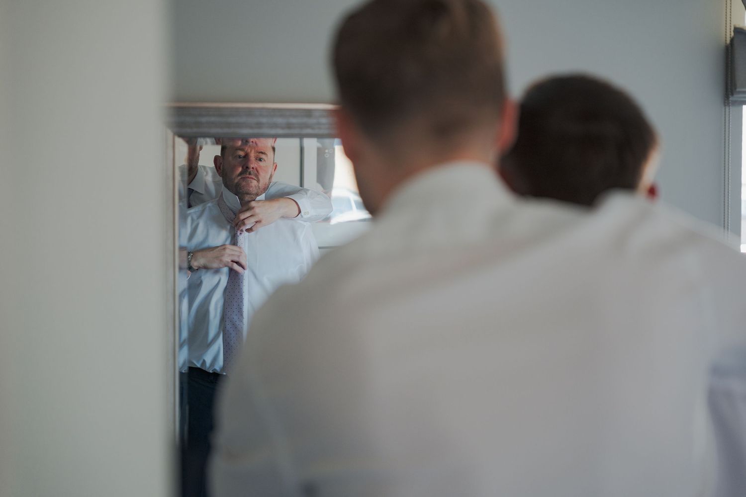 Someone adjusts their tie while looking in a mirror with blurred figures in the foreground.