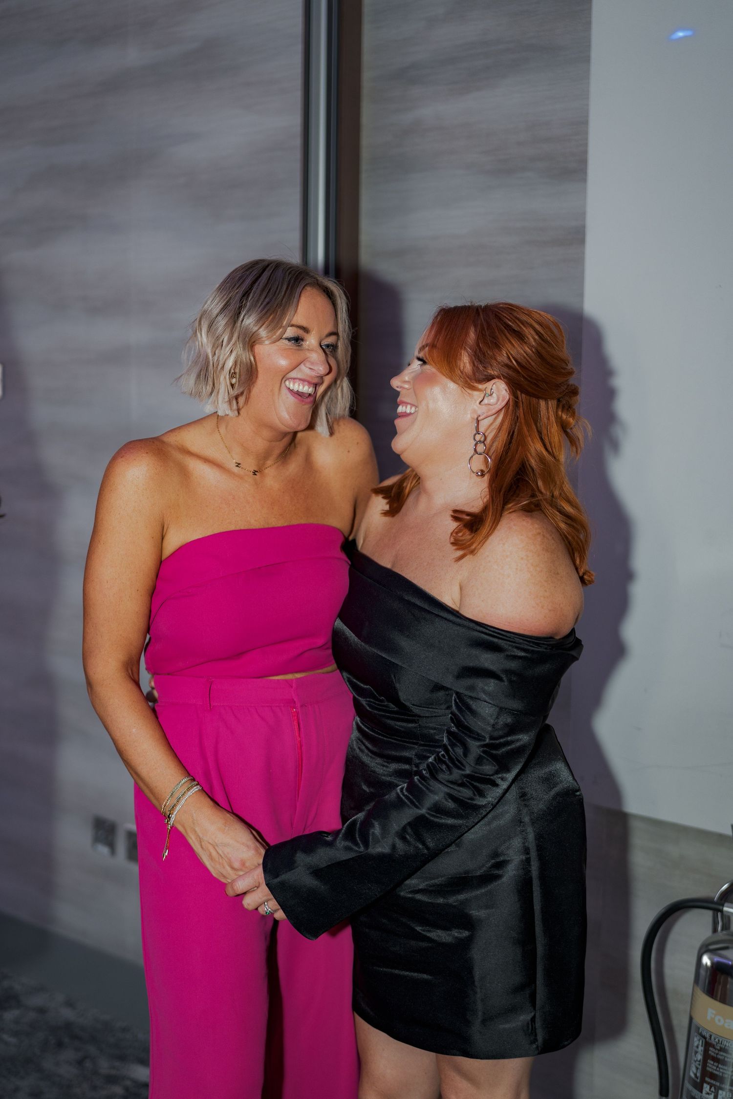 Two friends in formal dresses, one in pink and one in black, share a candid laughing moment at an event.