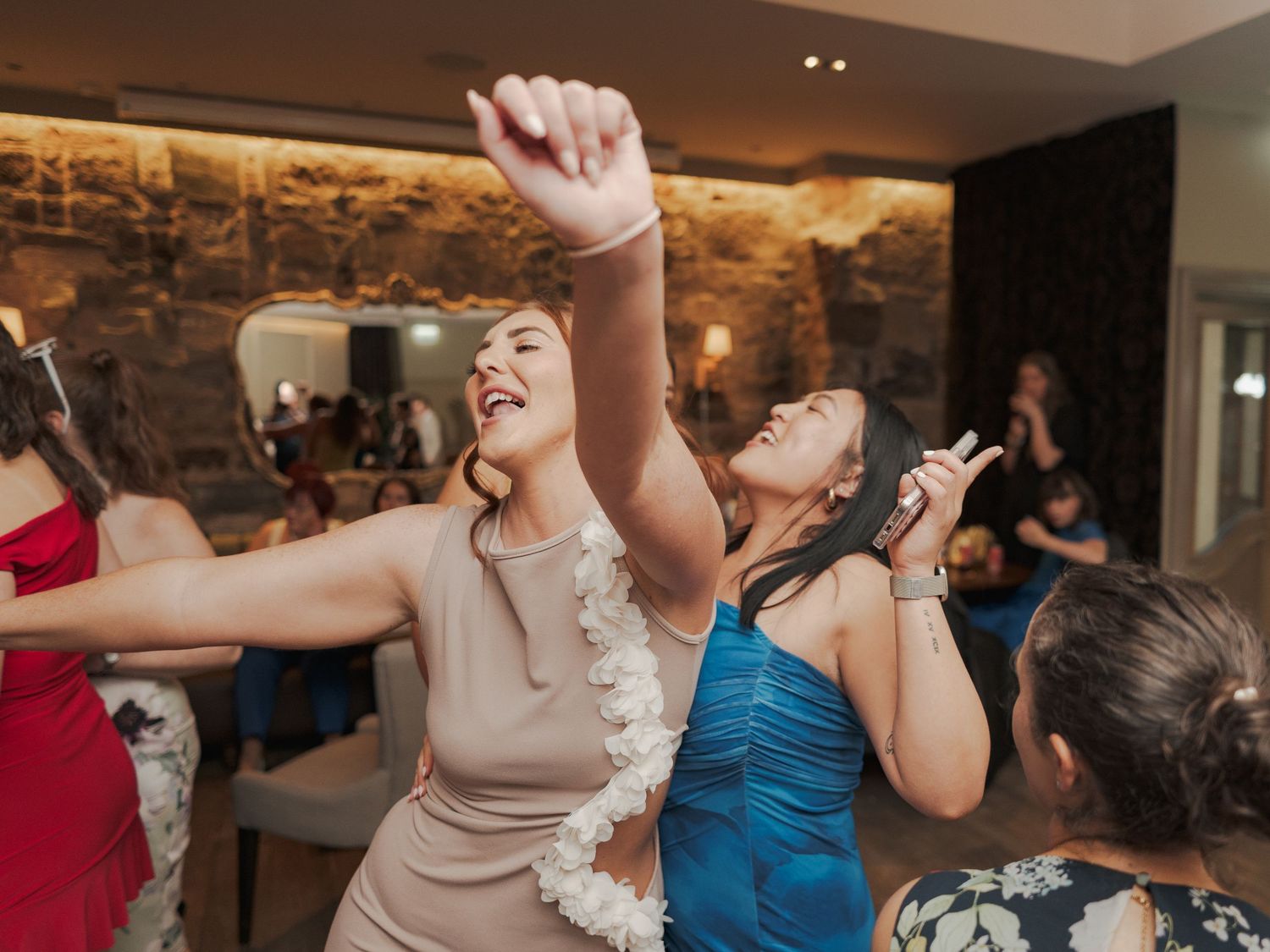 Wedding guests dance and celebrate with raised arms on a lively dance floor with stone walls in the background.