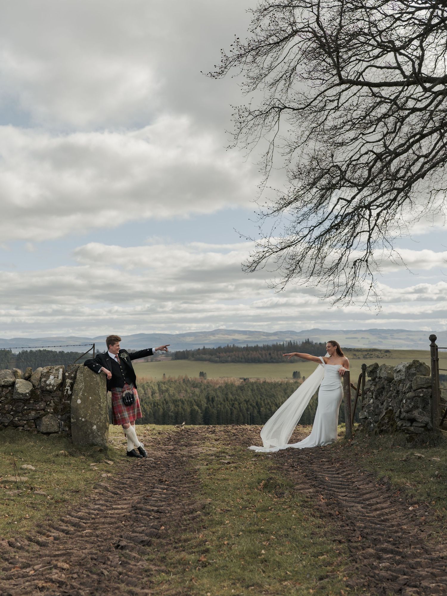 Scottish wedding couple in traditional kilt and bridal gown posing by stone wall with scenic Highland landscape backdrop.