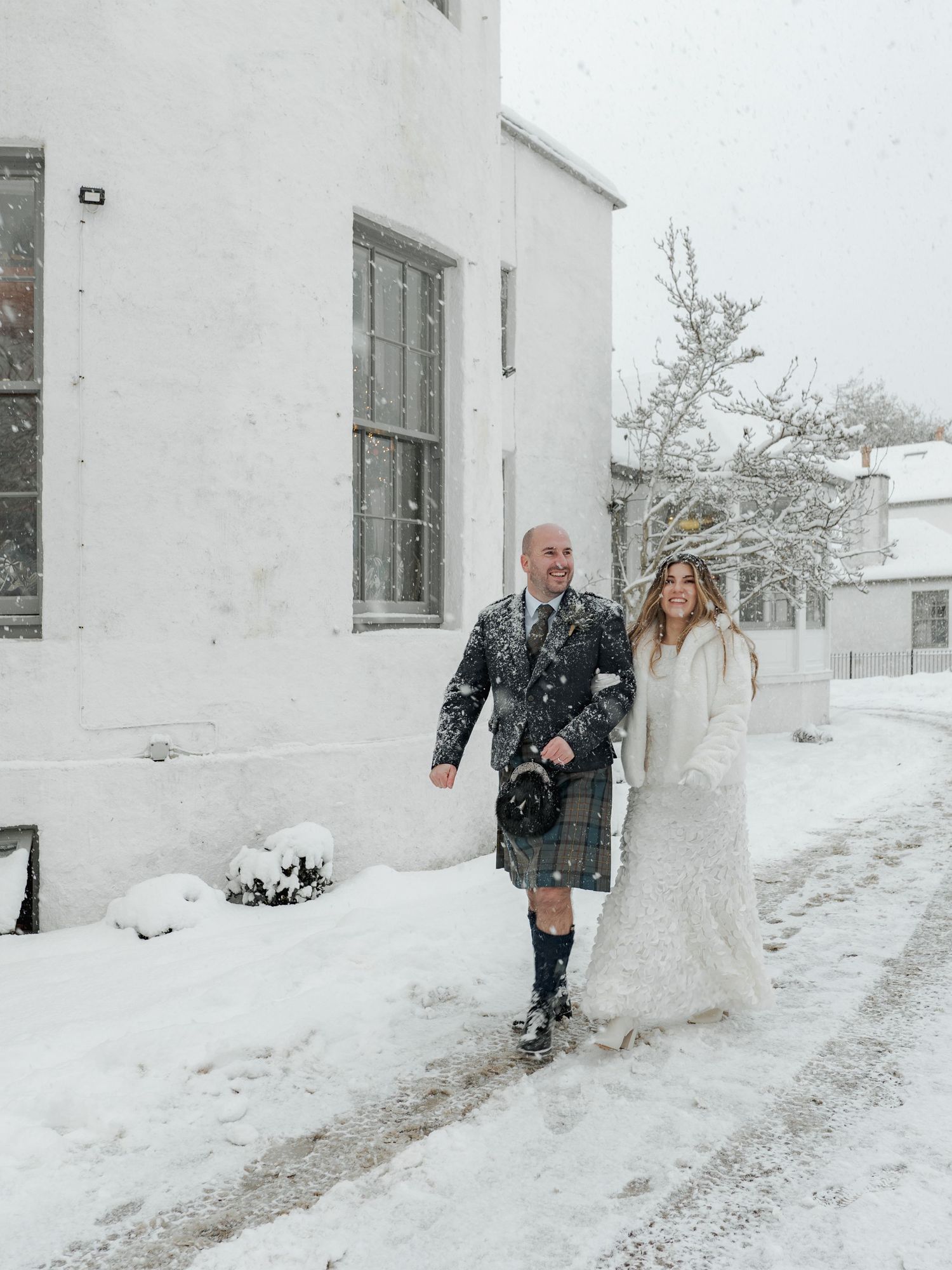 A couple in winter wedding attire walk together in the snow outside a white building with tall windows.