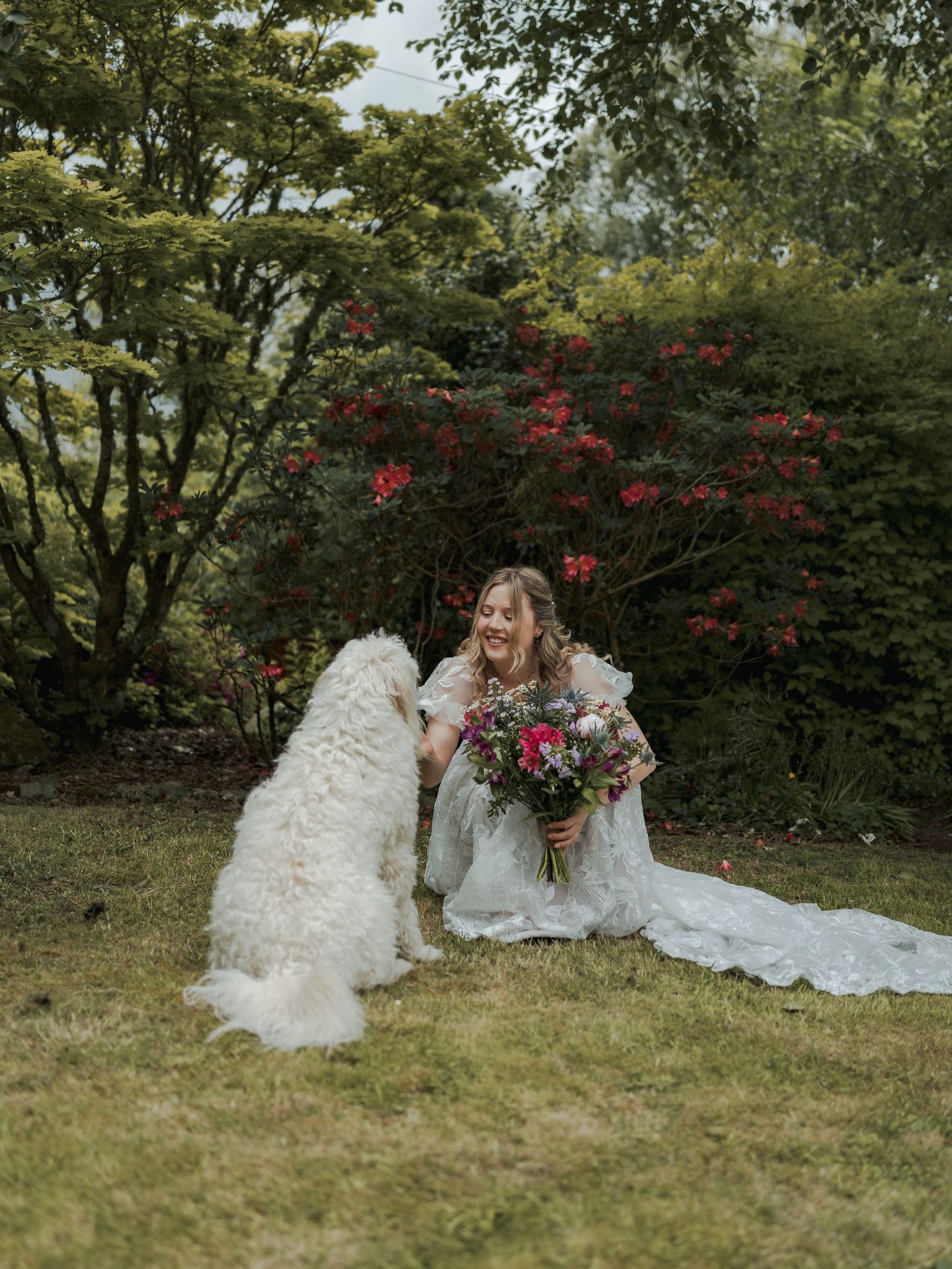 Bride in white gown sits on grass holding colorful bouquet while a fluffy white dog interacts with her in a lush garden.