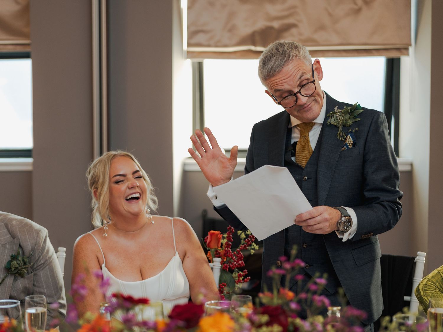 Wedding guests share joyful laughter during a reception celebration with warm lighting and floral decor.