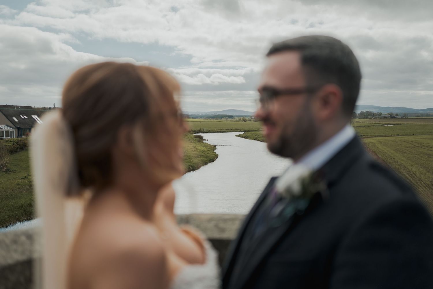 Bride and groom share a romantic moment outdoors with a scenic river and rolling hills in the background on their wedding day.