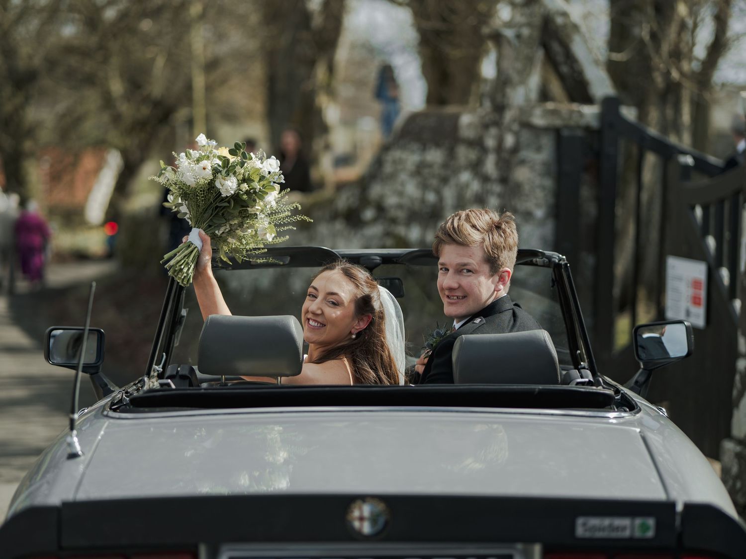 Newlywed couple smiling in a black convertible car, bride holding bouquet aloft, groom in suit, on a tree-lined street.
