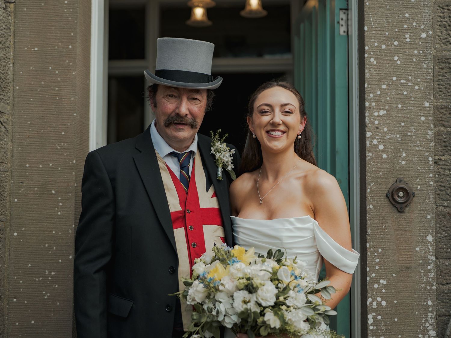 Bride in off-shoulder gown holding floral bouquet poses with man in dark suit and top hat with St George cross tie.