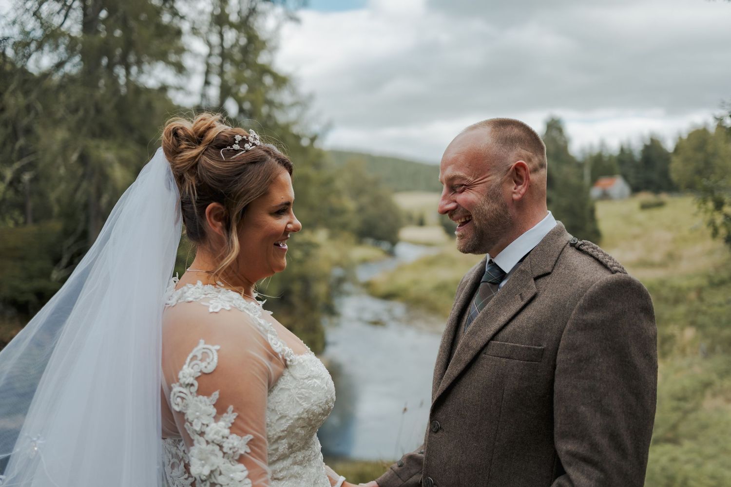 A bride and groom share a joyful moment outdoors in a scenic Scottish highland setting on their wedding day.
