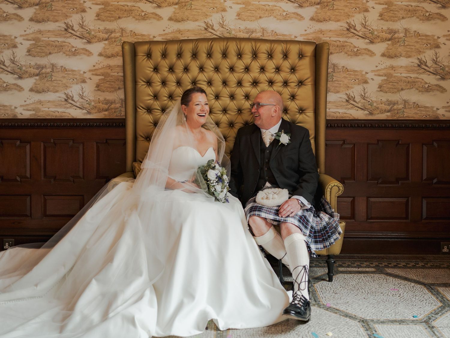 Bride in white gown and groom in Scottish kilt laughing together on a tufted gold settee at their wedding.