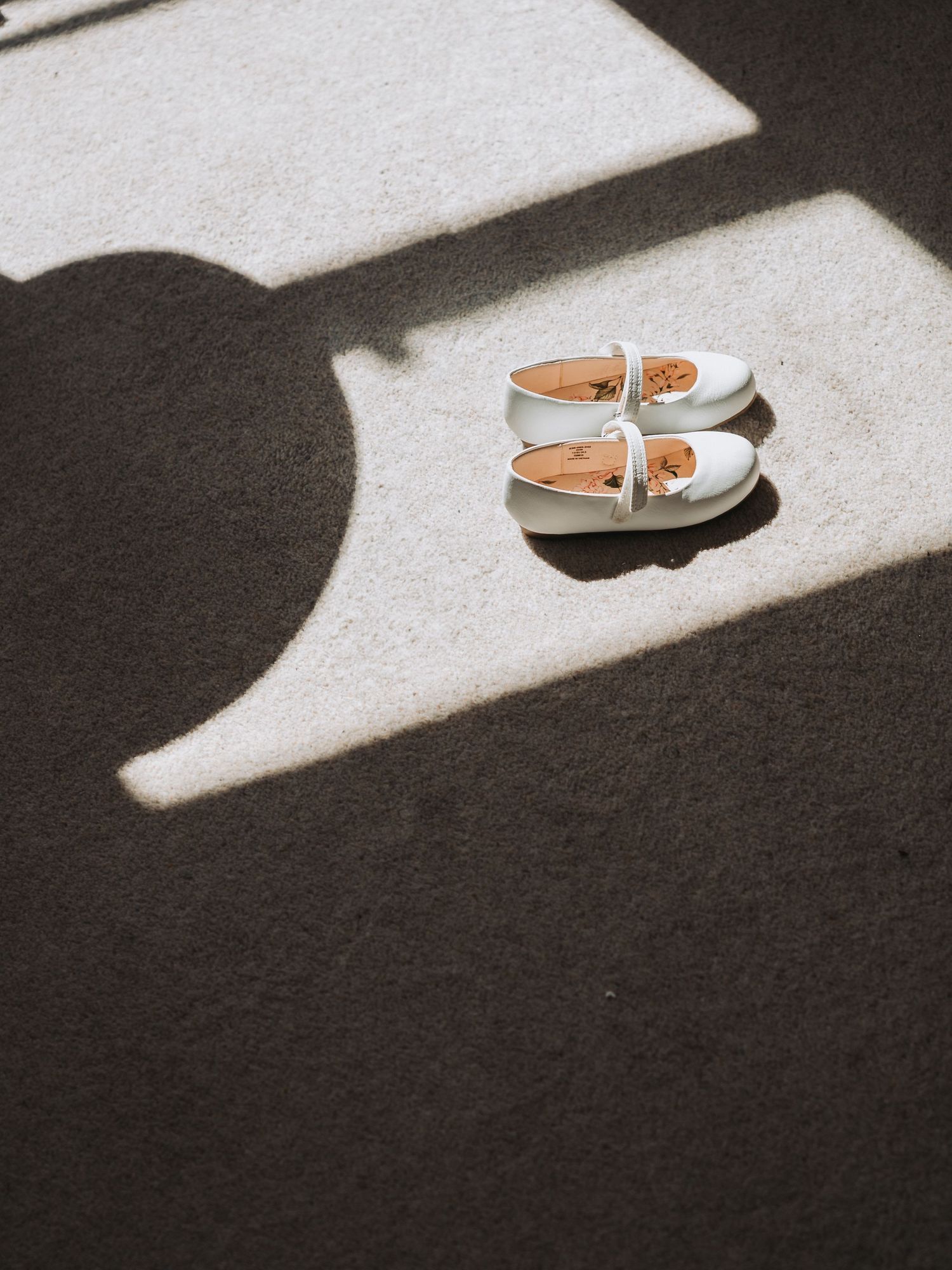 A pair of tan leather Mary Jane shoes casting dramatic shadows on a light carpet in contrasting lighting.