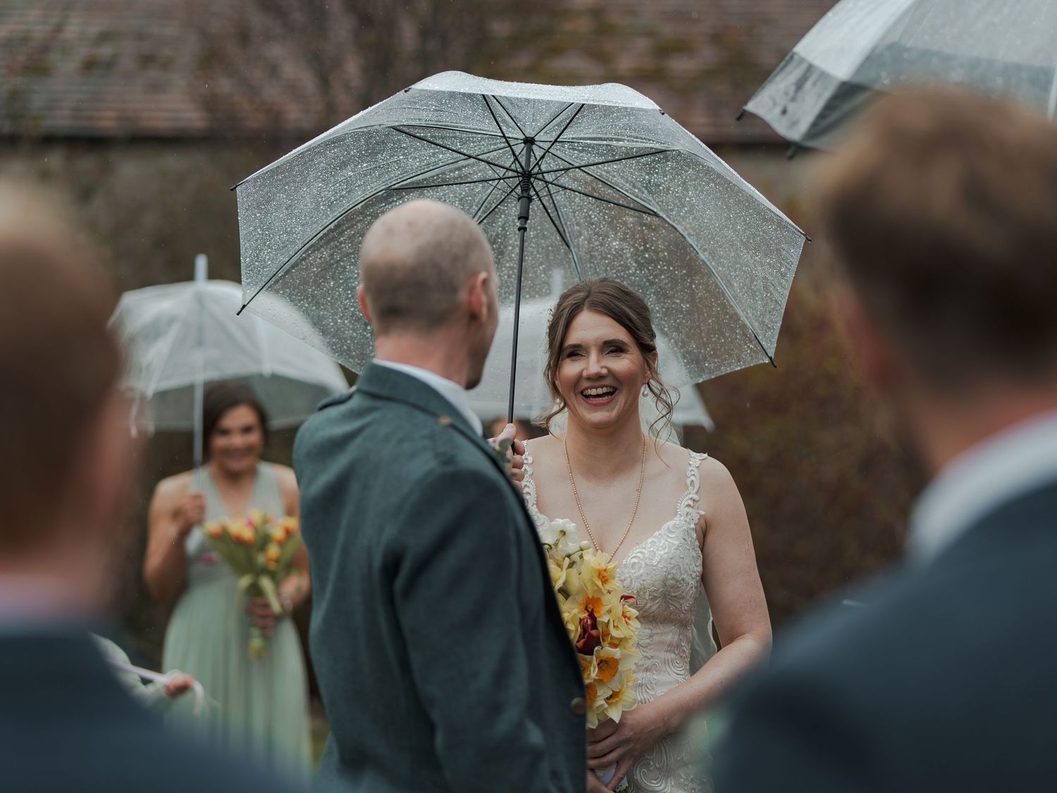 Smiling bride holding clear umbrella in rain, greeting guests with yellow bouquet at outdoor wedding ceremony.
