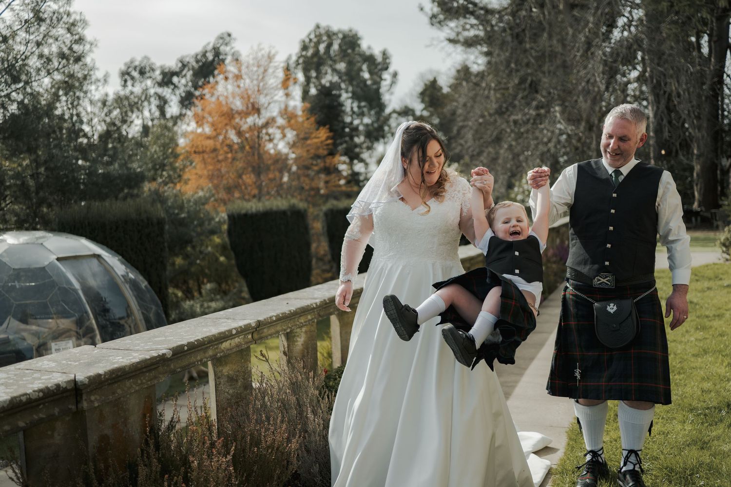 Bride in white gown and veil walks with man in Scottish kilt carrying a baby at an outdoor autumn wedding.