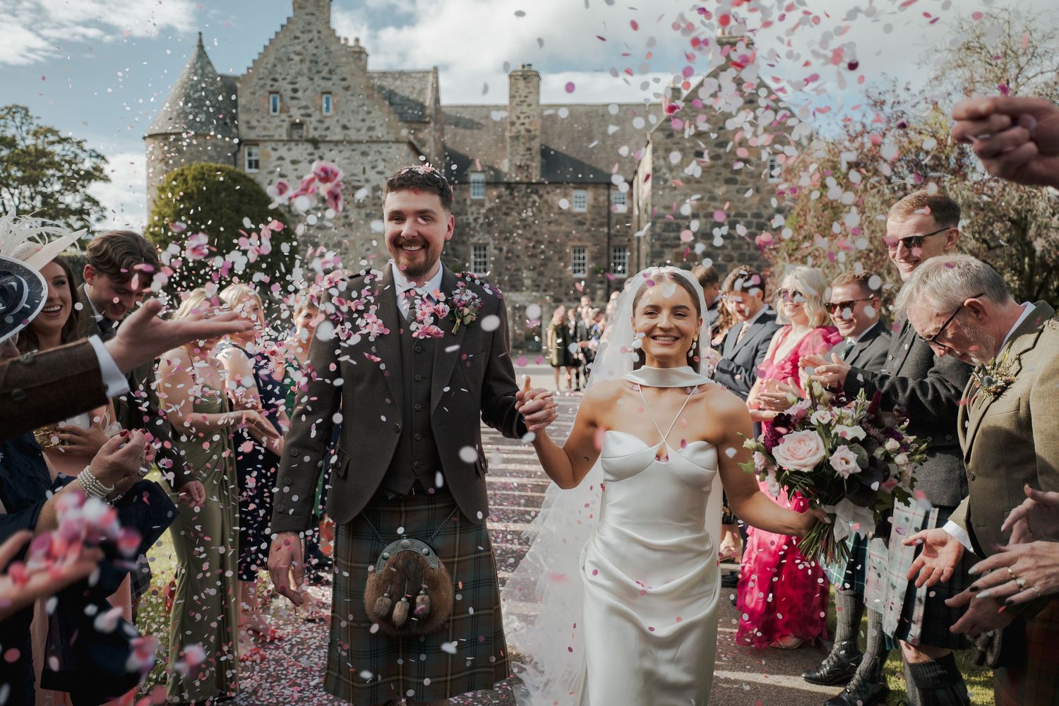 Bride and groom walk through confetti shower outside historic Scottish venue, groom in kilt, bride in strapless gown holding bouquet.