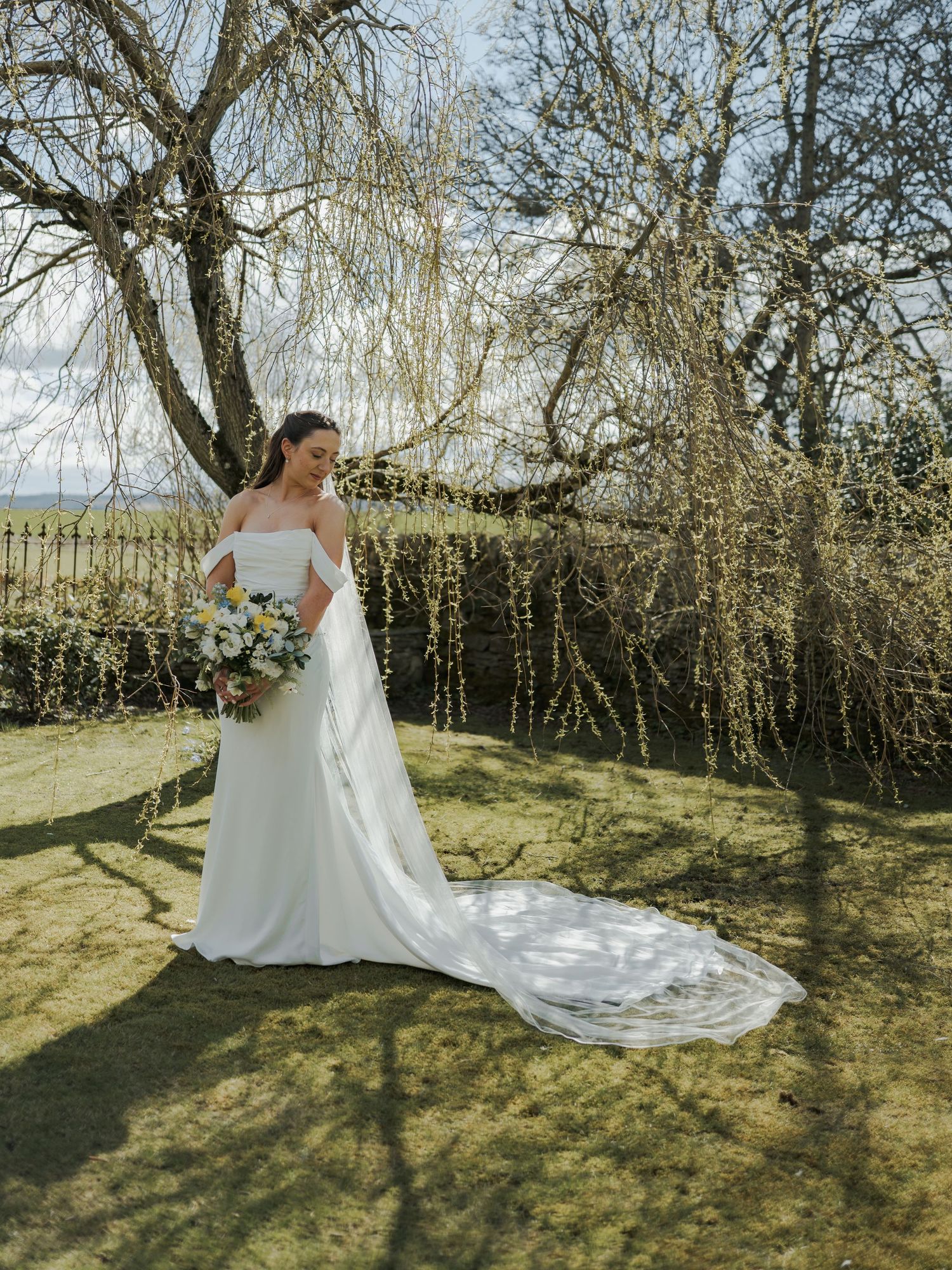 Bride in off-shoulder white gown with long train holds bouquet beside bare winter trees in garden.
