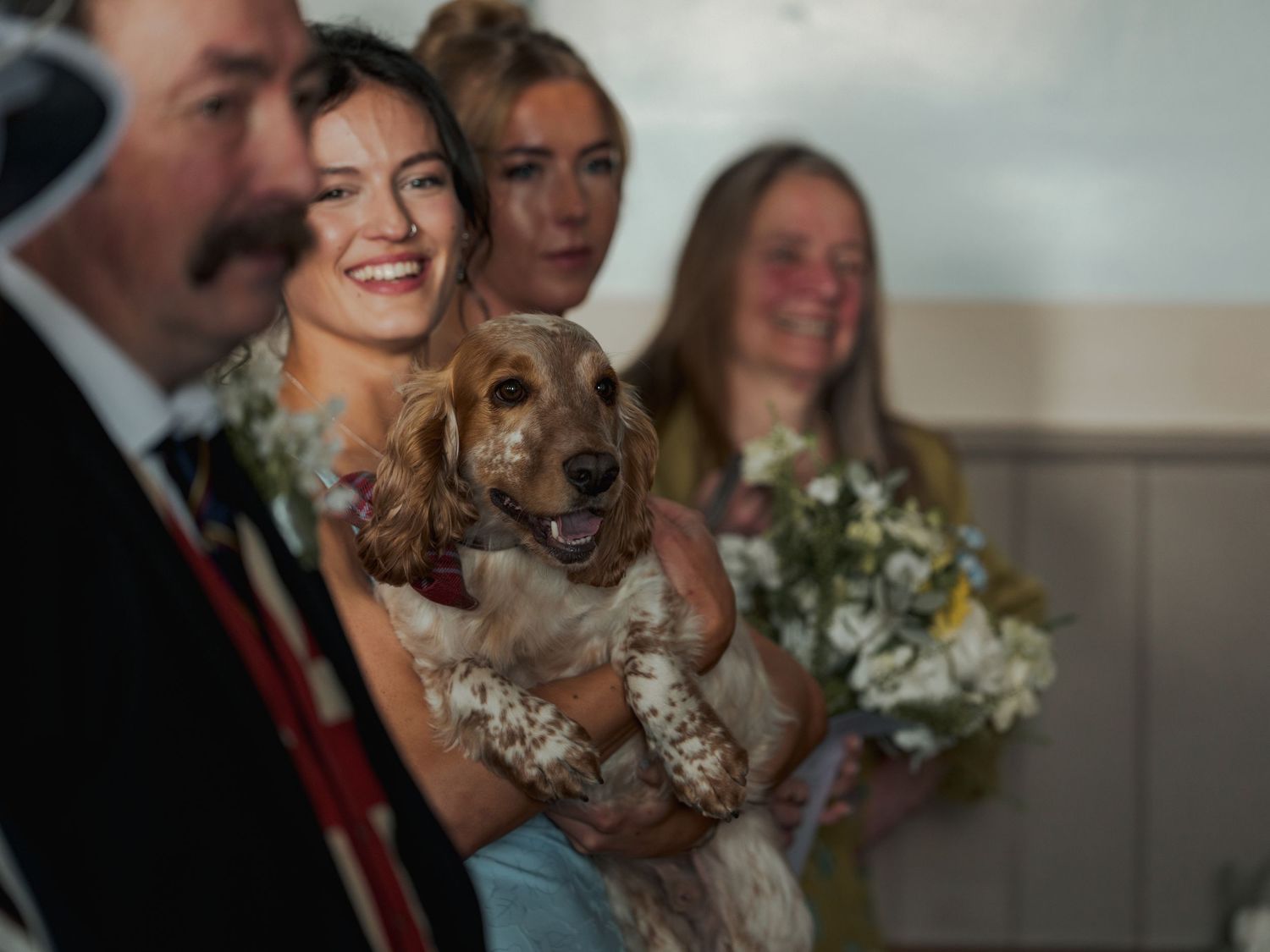 Bridesmaid holding a golden cocker spaniel dog at a wedding ceremony with bouquets and guests in background.