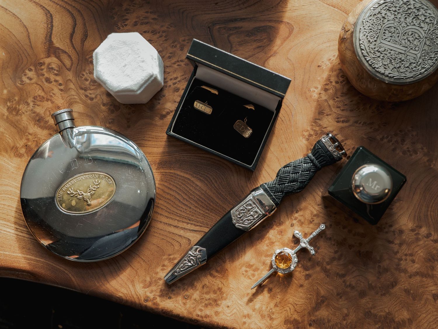 Scottish accessories flat lay featuring a flask, sgian dubh, cufflinks, brooch, and decorative boxes on a wooden surface.