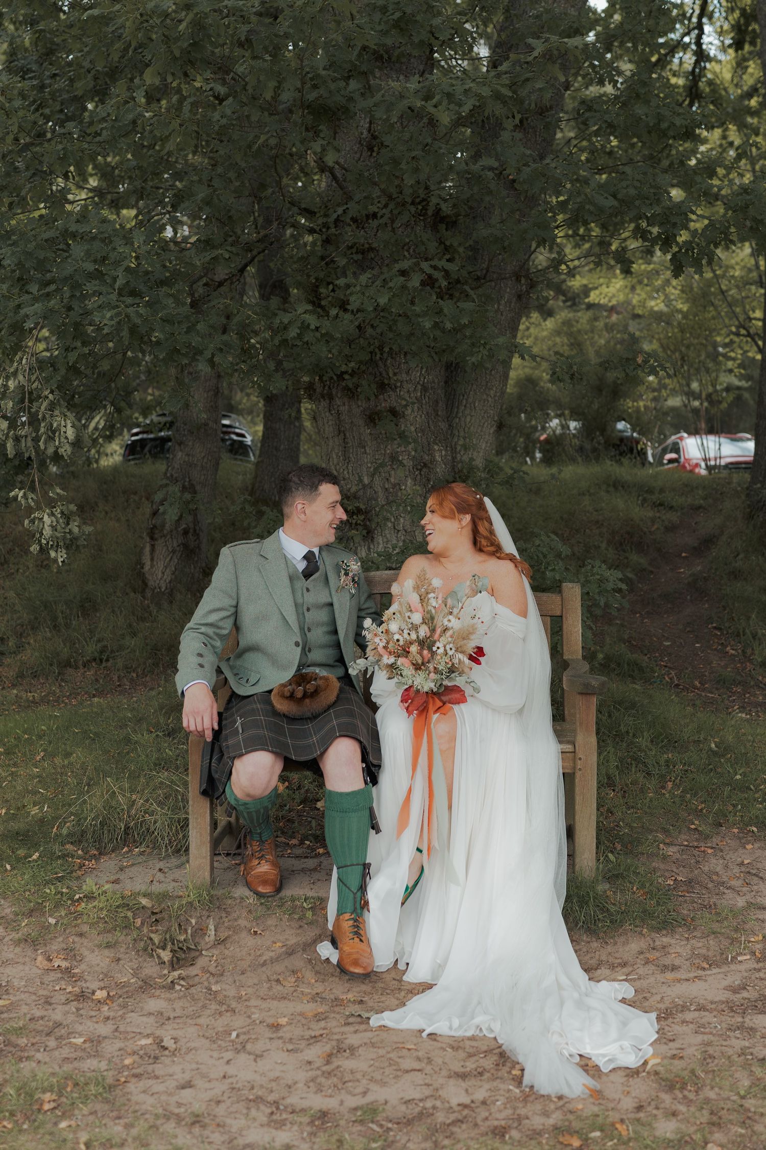 Couple sits on wooden bench in outdoor garden setting wearing elegant wedding attire at Banchory Lodge.