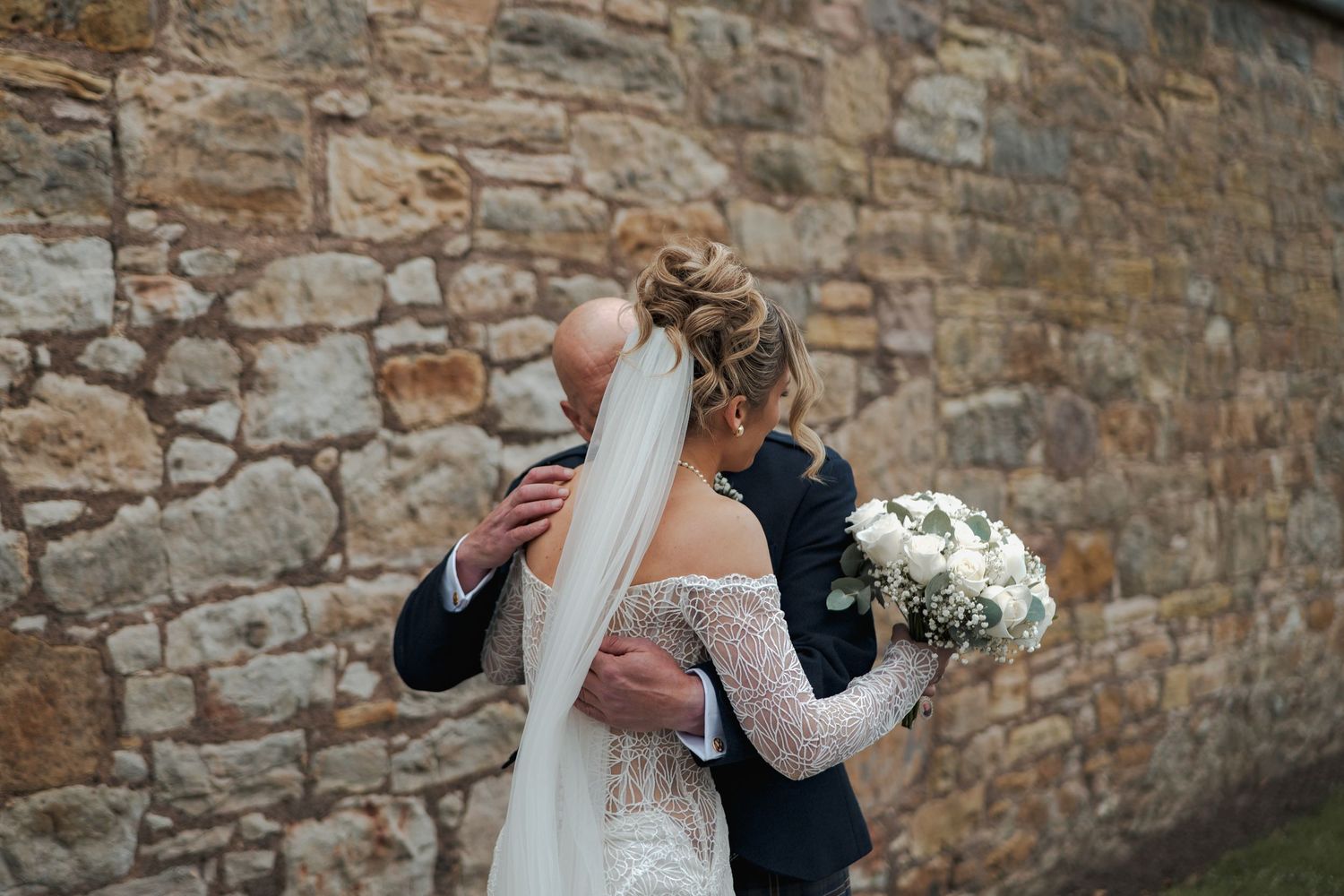 Newlyweds share an intimate moment against a rustic stone wall while holding a white floral bouquet.