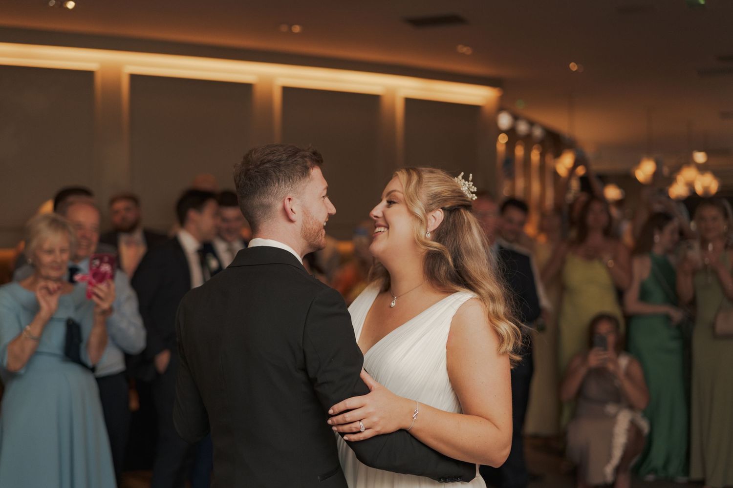 Wedding guests dance together on a dimly lit ballroom floor during an evening reception celebration.