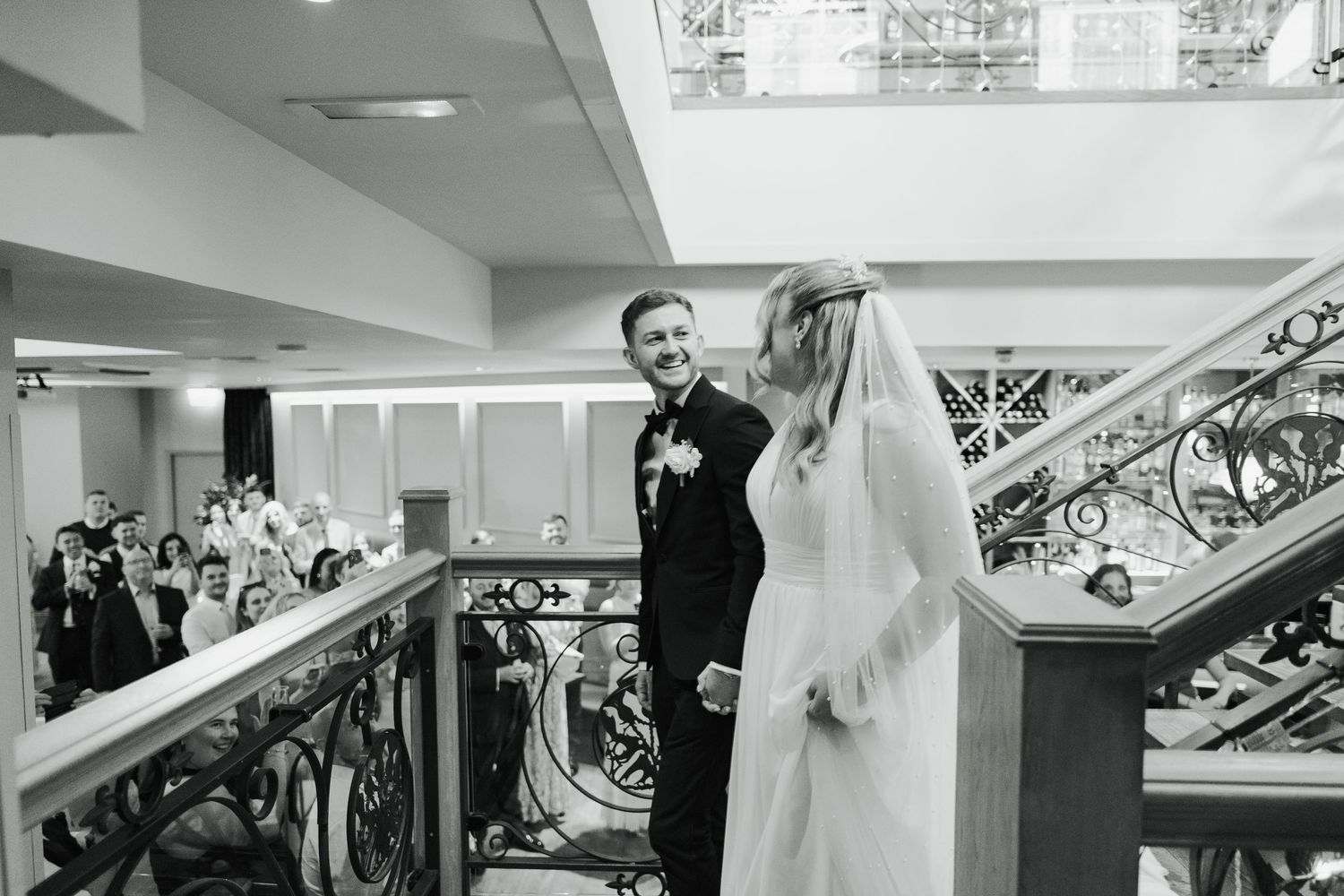 A bride in a flowing white wedding dress walks down an elegant staircase with ornate railings and decorative details.