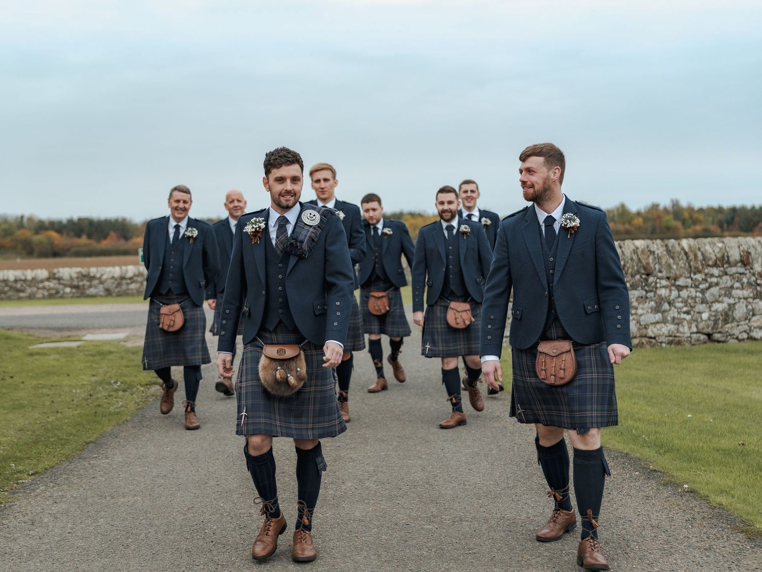 Groomsmen in black suits and kilts posing together outdoors on a cloudy day.