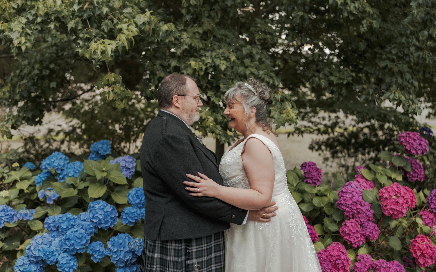 Couple in wedding attire embrace outdoors surrounded by colorful blue and pink hydrangeas.