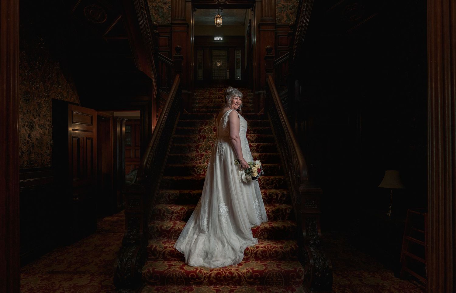 Bride in white gown stands on ornate staircase in elegant dark wood manor, holding bouquet.
