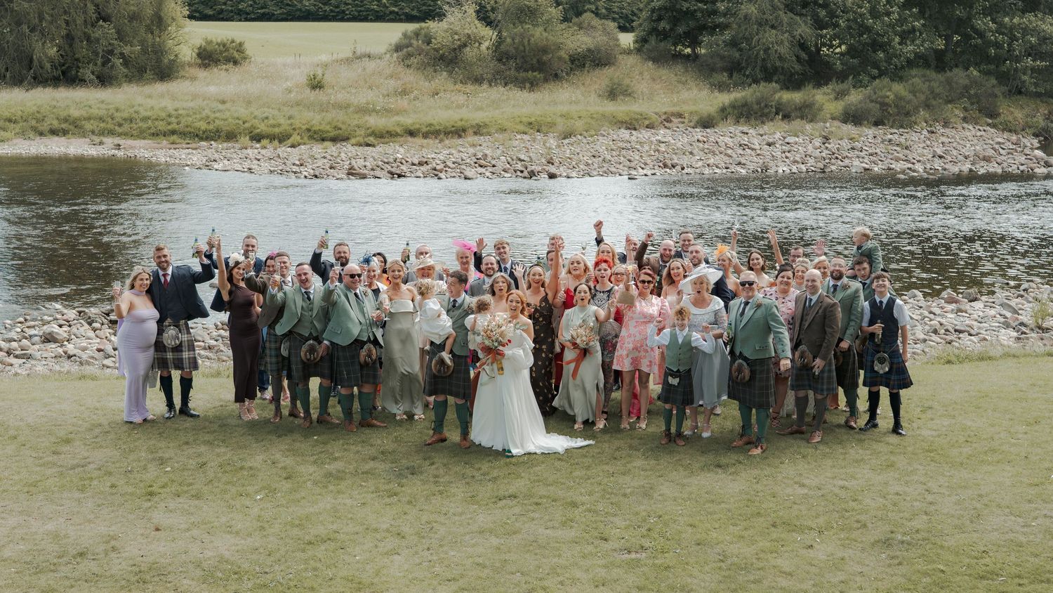 Wedding party lined up in panoramic view by a scenic riverbank and rocky shoreline during an outdoor celebration.