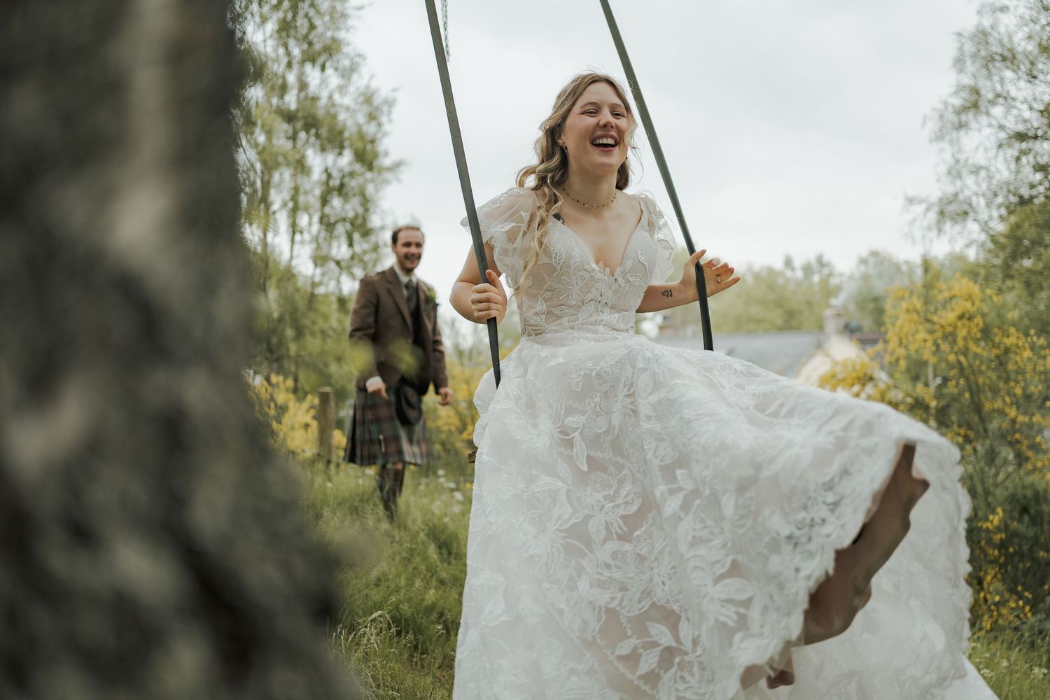 Bride in lace gown laughing on a swing while groom in tartan kilt pushes her outdoors at a woodland wedding.