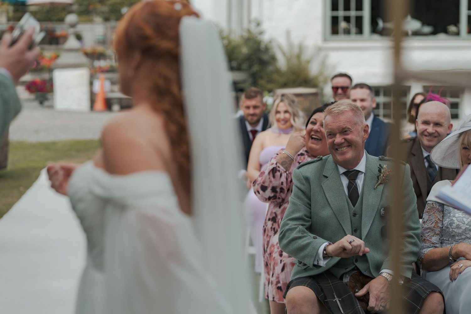 During an outdoor wedding ceremony, a person in a green suit holds up a ring in a tender moment.